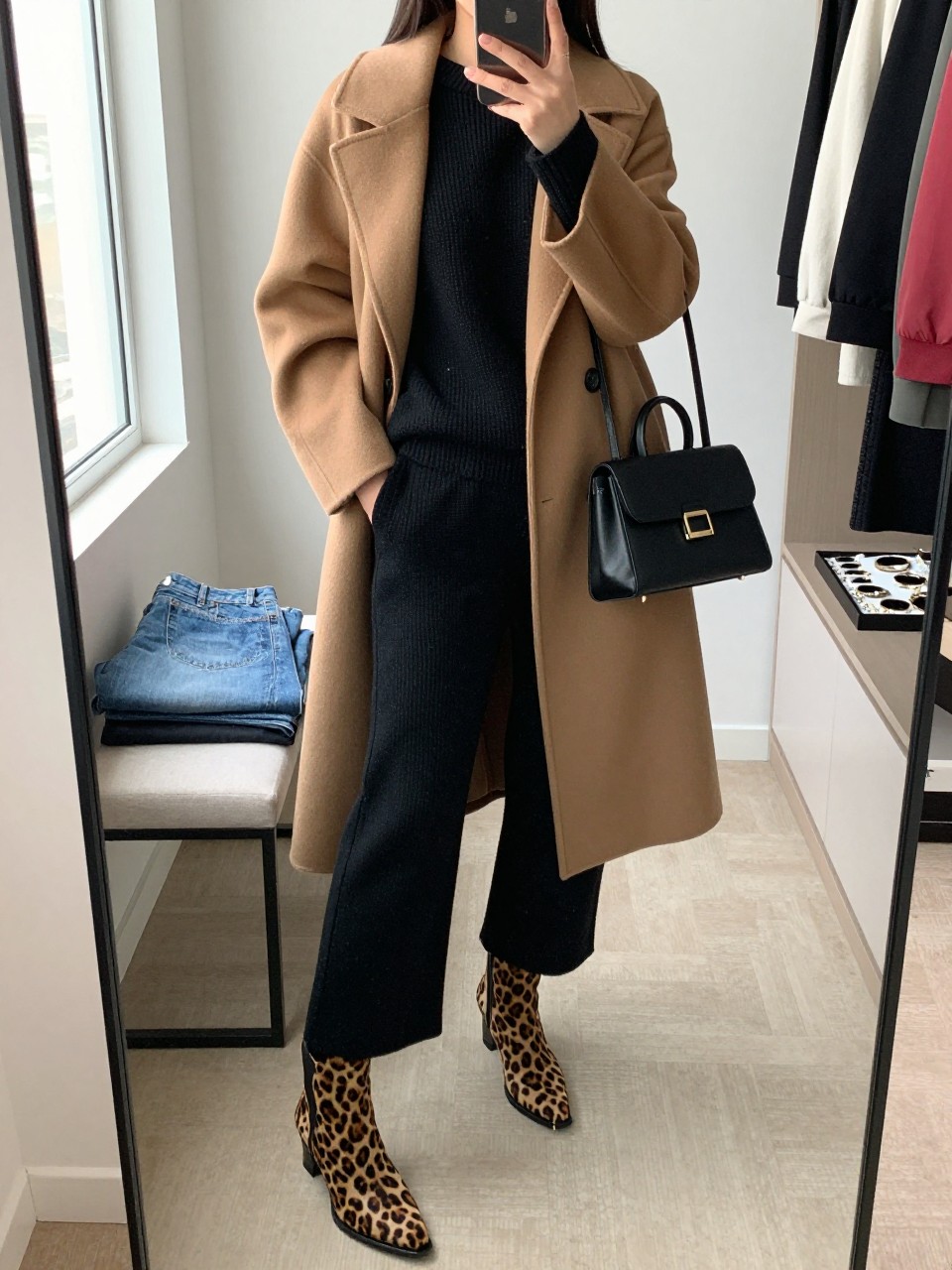 mirror selfie of a woman wearing the exact outfit described above, clearly showing animal print calf-hair ankle boots in brown leopard print, a black knit sweater, matching black knit wide-leg pants, a camel long coat, a black mini handbag, and small gold hoops, holding a phone in front of a mirror, cropped at NECK so the face does not show, with a relaxed casual selfie pose, in a walk-in closet mirror, natural window light, with folded denim jeans on a bench and a jewelry tray on the vanity, latest iPhone photo quality.