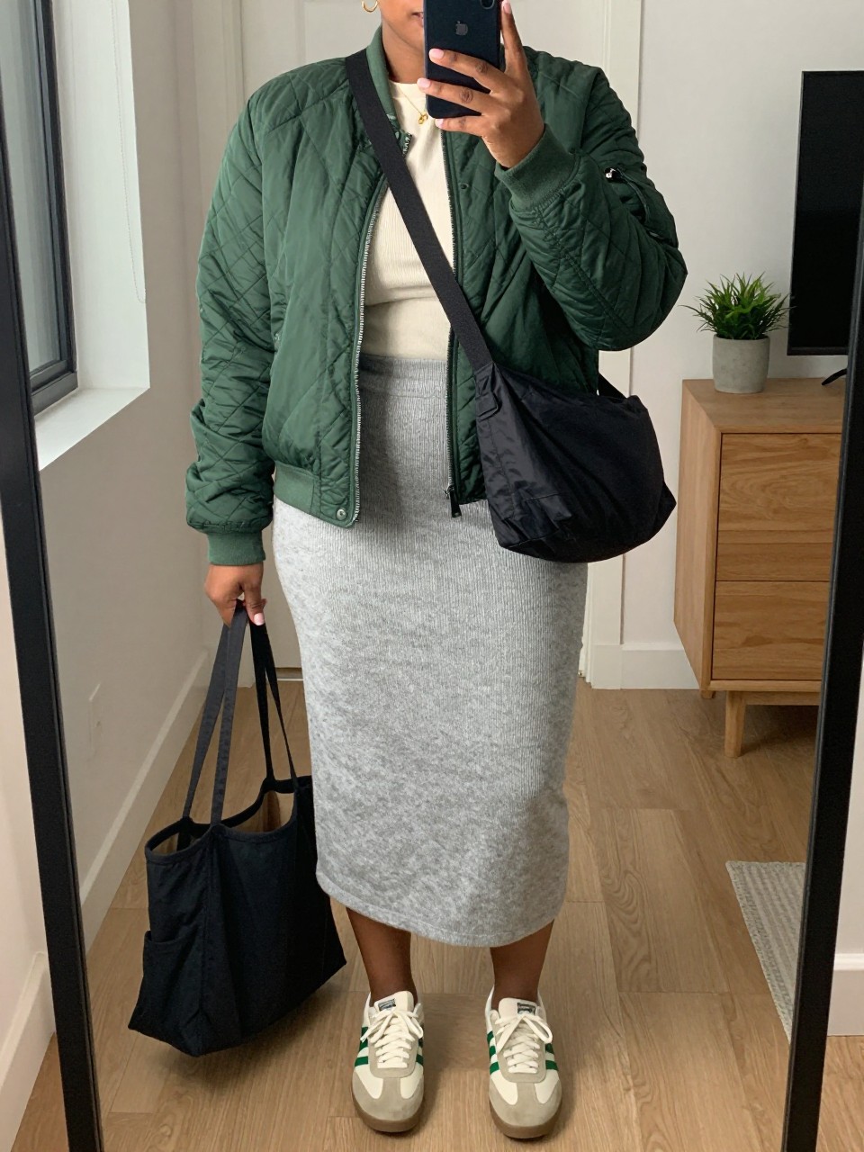 mirror selfie of a plus size Black woman wearing the exact outfit described above, a forest green quilted bomber jacket over a cream fine knit long-sleeve top, heather gray knit midi skirt, cream and green retro sneakers, and black nylon crossbody bag, clearly showing the same clothing items, colors, fabrics, and accessories listed in the Outfit Pieces section, holding a phone in front of a mirror, cropped at NECK so the face does not show, with a relaxed casual selfie pose, in a stylish city apartment, soft indoor lighting, including a tote bag resting on the floor and a small plant on a dresser, latest iPhone photo quality.