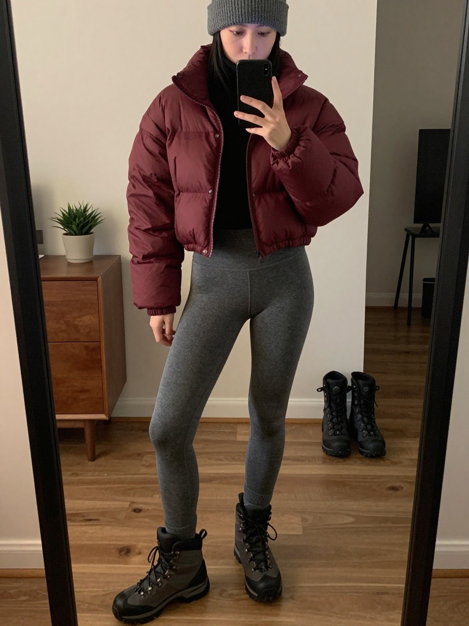 mirror selfie of a woman wearing the exact outfit described above, clearly showing a burgundy cropped puffer jacket, soft gray thermal leggings, black merino wool turtleneck, black hiking boots, and a gray knit beanie, holding a phone in front of a mirror, cropped at NECK so the face does not show, with a relaxed casual selfie pose, in a stylish city apartment, warm moody lighting, with a small plant on a dresser and a pair of boots on the floor nearby, latest iPhone photo quality.