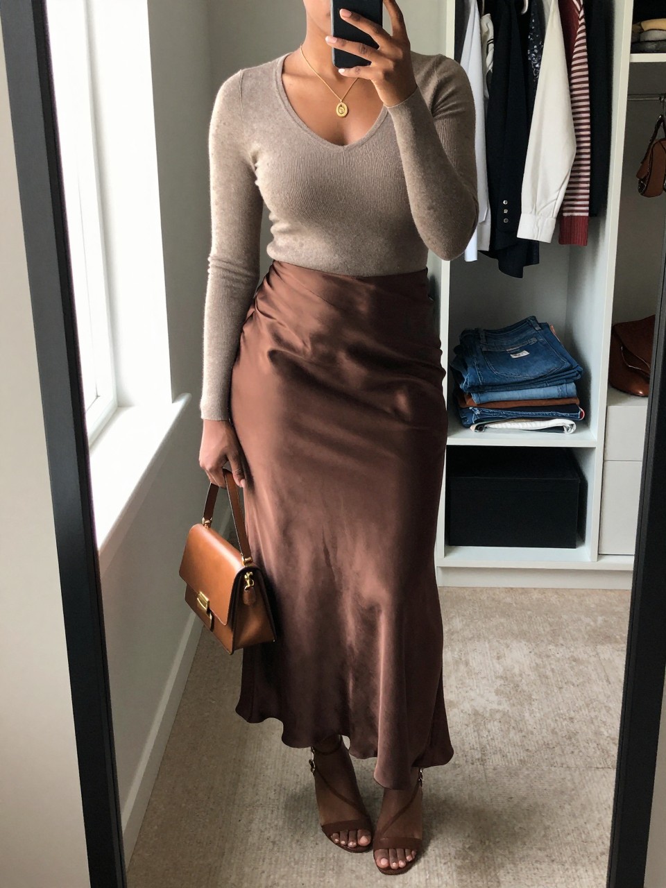 mirror selfie of a Black woman wearing the exact outfit described above, clearly showing a taupe fitted knit top, chocolate satin maxi skirt, brown strappy heels, gold pendant necklace, and a dark tan mini bag, holding a phone in front of a mirror, cropped at NECK so the face does not show, with a relaxed casual selfie pose, in a walk-in closet mirror, natural window light, with an open closet with clothes visible and folded denim jeans on a bench, latest iPhone photo quality.