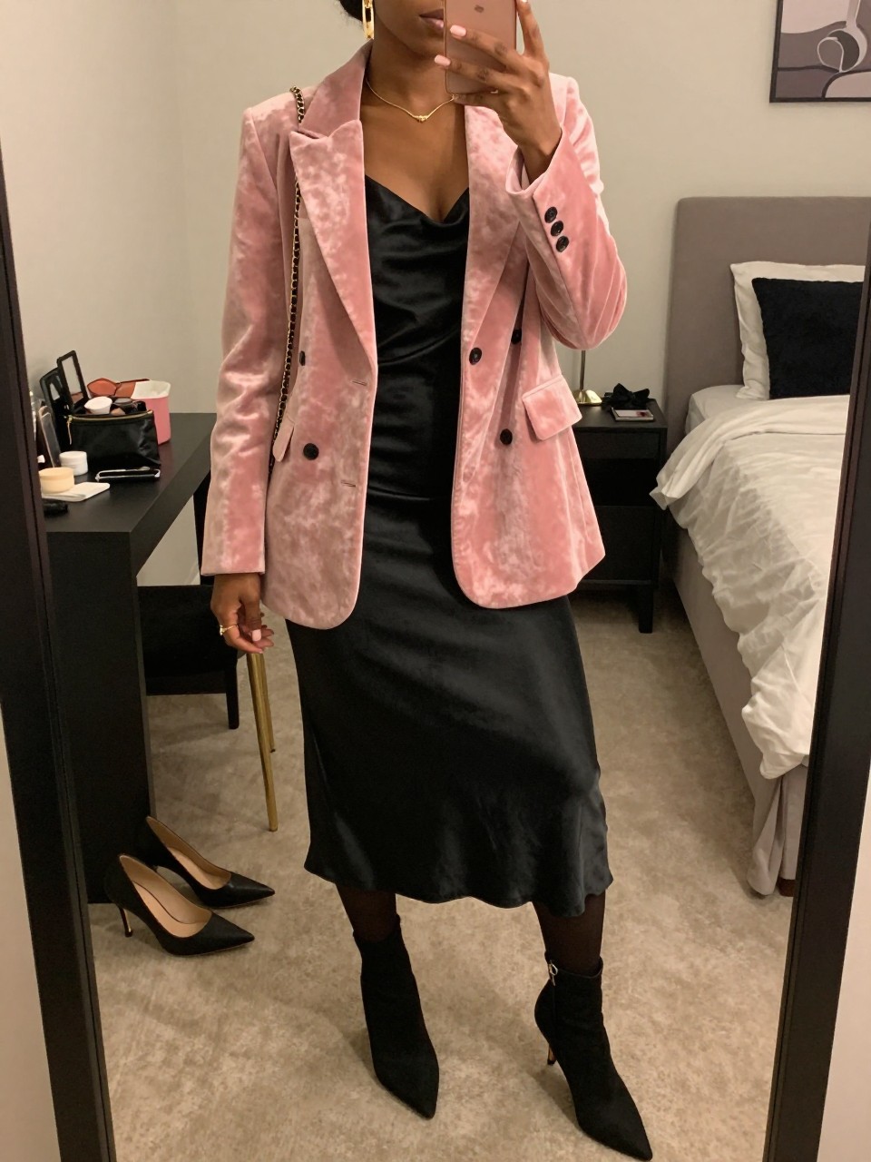 mirror selfie of a Black woman wearing a rose pink velvet blazer over a black satin midi slip dress, sheer black tights, black pointed kitten-heel ankle boots, gold drop earrings, and a small black chain-strap bag, clearly showing the same clothing items, colors, fabrics, and accessories listed in the Outfit Pieces section, holding a phone in front of a mirror, cropped at NECK so the face does not show, with a relaxed casual selfie pose, in a feminine-themed bedroom, warm moody lighting, including a pair of heels on the floor nearby and a cosmetic bag on the counter, latest iPhone photo quality.