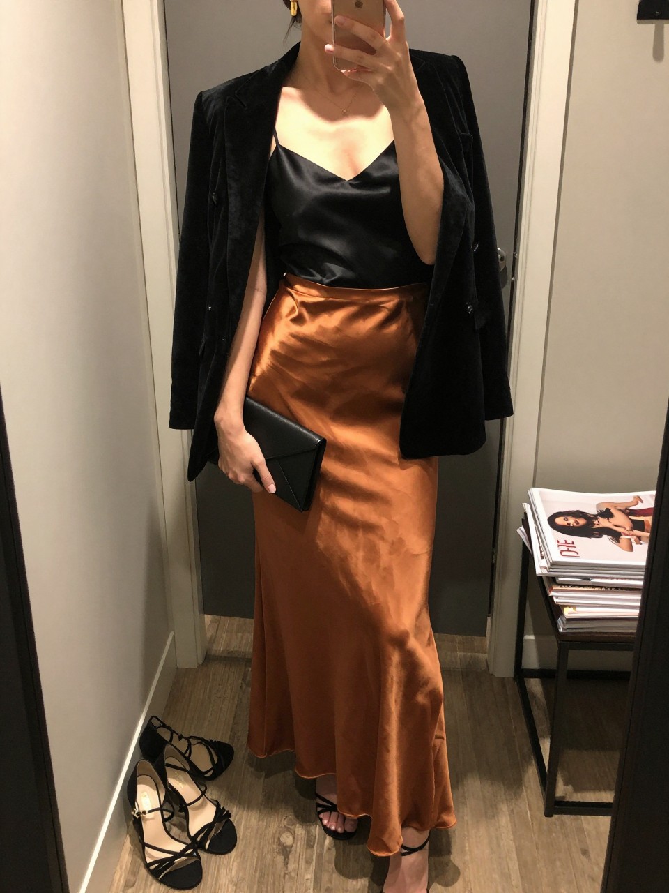 mirror selfie of a woman wearing the exact outfit described above, clearly showing a copper satin maxi skirt, fitted black satin cami, black velvet blazer, black strappy heels, black envelope clutch, and gold drop earrings, holding a phone in front of a mirror, cropped at NECK so the face does not show, with a relaxed casual selfie pose, in a walk-in closet mirror, warm moody lighting, with a pair of heels on the floor nearby and a stack of fashion magazines on a side table, latest iPhone photo quality.