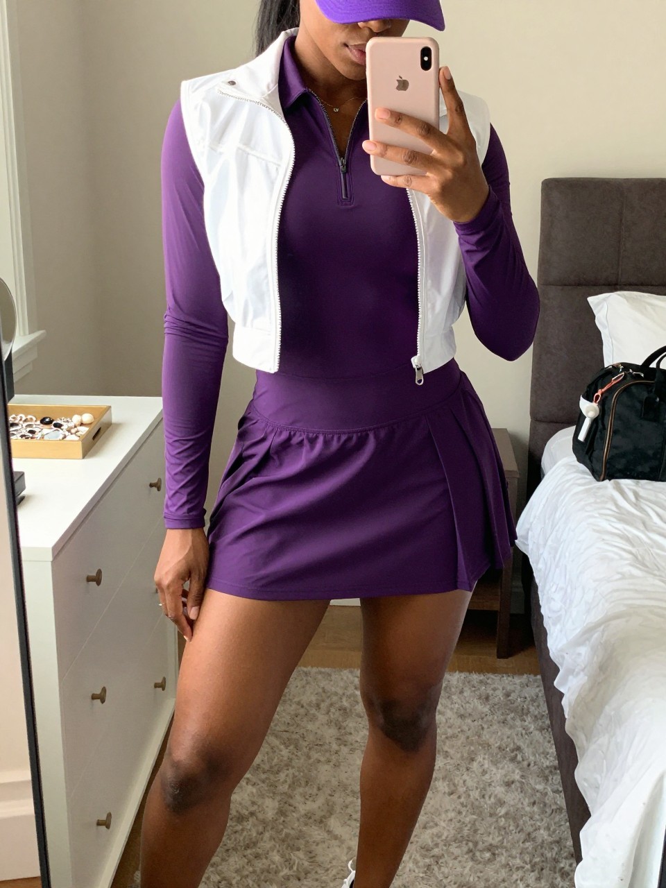 Mirror selfie of a Black woman with rich dark skin wearing a fitted plum-purple long-sleeve golf dress with a zip collar, pleated skirt, and built-in shorts, a cropped white performance jacket worn open, white spikeless golf sneakers, and a plum visor, clearly showing the exact clothing items, colors, fabrics, and accessories listed, holding a latest-model iPhone in front of a mirror, cropped at the neck so the face does not show, relaxed casual selfie pose, in a feminine-themed bedroom with soft indoor lighting, a jewelry tray on a vanity and a small golf duffel beside the bed, natural unstaged composition, latest iPhone photo quality.