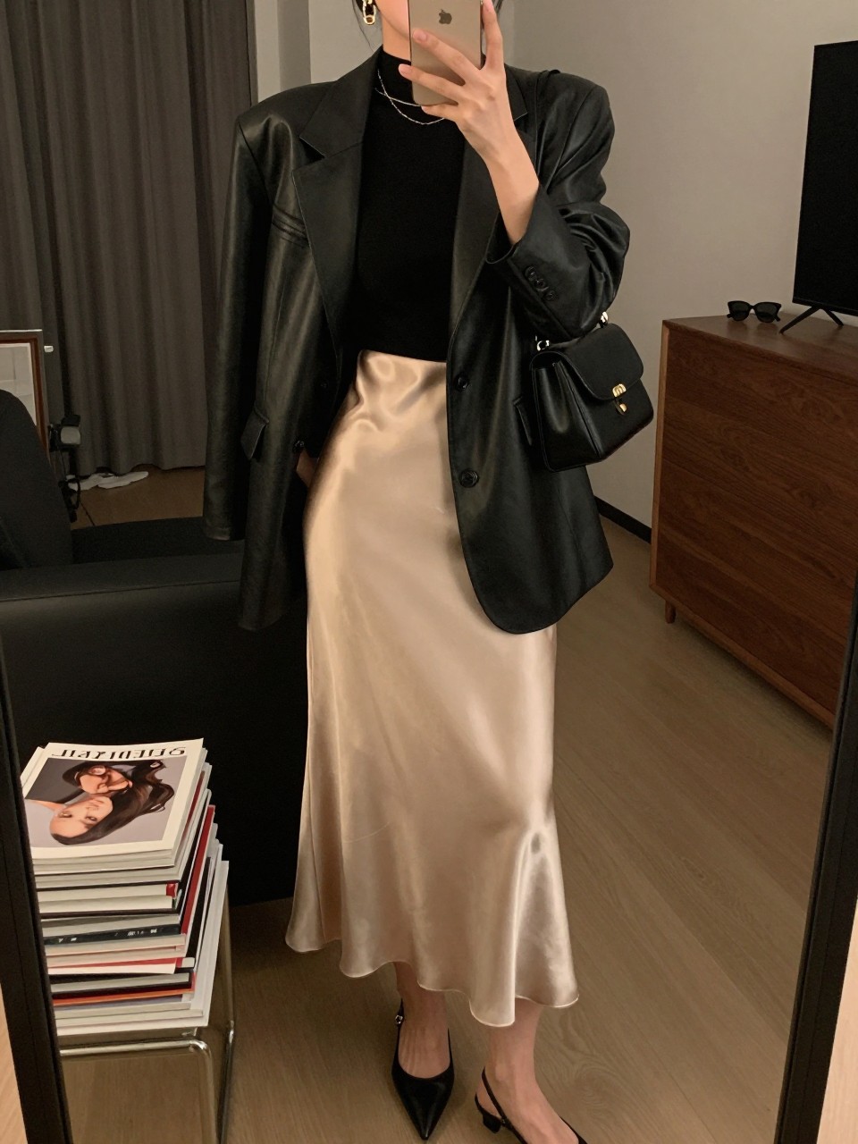 mirror selfie of a woman wearing the exact outfit described above, clearly showing a black leather blazer layered over a fitted black mock-neck top, champagne satin midi slip skirt, black slingback heels, a black mini shoulder bag, and gold drop earrings, holding a phone in front of a mirror, cropped at NECK so the face does not show, with a relaxed casual selfie pose, in a stylish city apartment, warm moody lighting, with a stack of fashion magazines on a side table and sunglasses placed on a dresser, latest iPhone photo quality.
