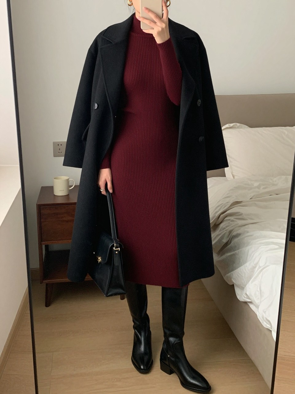 mirror selfie of a woman wearing the exact outfit described above, clearly showing a burgundy ribbed knit midi dress, tall black leather boots, a black wool coat draped open, gold hoop earrings, and a black structured handbag, holding a phone in front of a mirror, cropped at NECK so the face does not show, with a relaxed casual selfie pose, in a cozy bedroom mirror setup, soft indoor lighting, with a coffee mug on a bedside table and soft pillows on an unmade bed, latest iPhone photo quality.