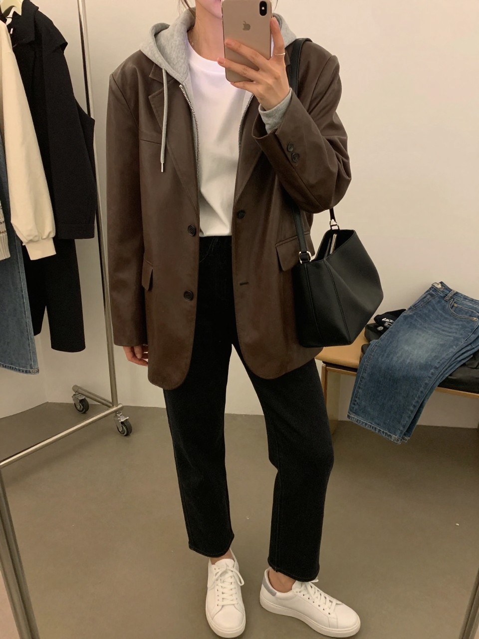 Mirror selfie of a woman wearing an oversized chocolate brown leather blazer layered over a heather gray cotton hoodie and a fitted white crew-neck T-shirt, black straight-leg jeans, clean white leather sneakers, and a structured black shoulder bag, clearly showing the exact clothing items, colors, fabrics, and accessories listed, holding a latest-model iPhone in front of a mirror, cropped at the neck so the face does not show, relaxed casual selfie pose, in a fashion studio mirror corner with warm moody lighting, a clothing rack with garments behind and folded denim jeans on a bench, natural casual composition, latest iPhone photo quality.