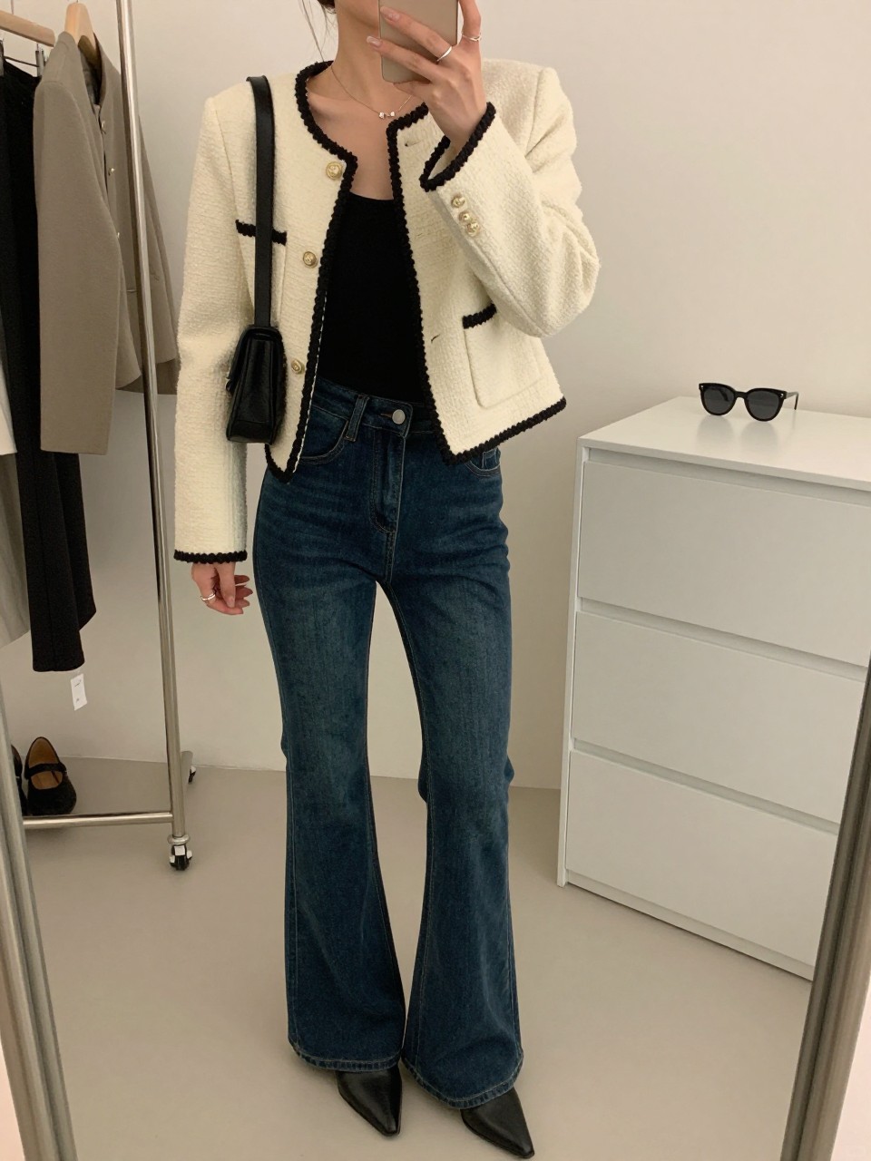 mirror selfie of a woman wearing a cream bouclé jacket with black trim, fitted black scoop-neck top, dark indigo flare jeans, black pointed ankle boots, and a quilted black shoulder bag, clearly showing the same clothing items, colors, fabrics, and accessories listed in the Outfit Pieces section, holding a phone in front of a mirror, cropped at NECK so the face does not show, with a relaxed casual selfie pose, in a fashion studio mirror corner, soft indoor lighting, including a clothing rack with garments behind and sunglasses placed on a dresser, latest iPhone photo quality.