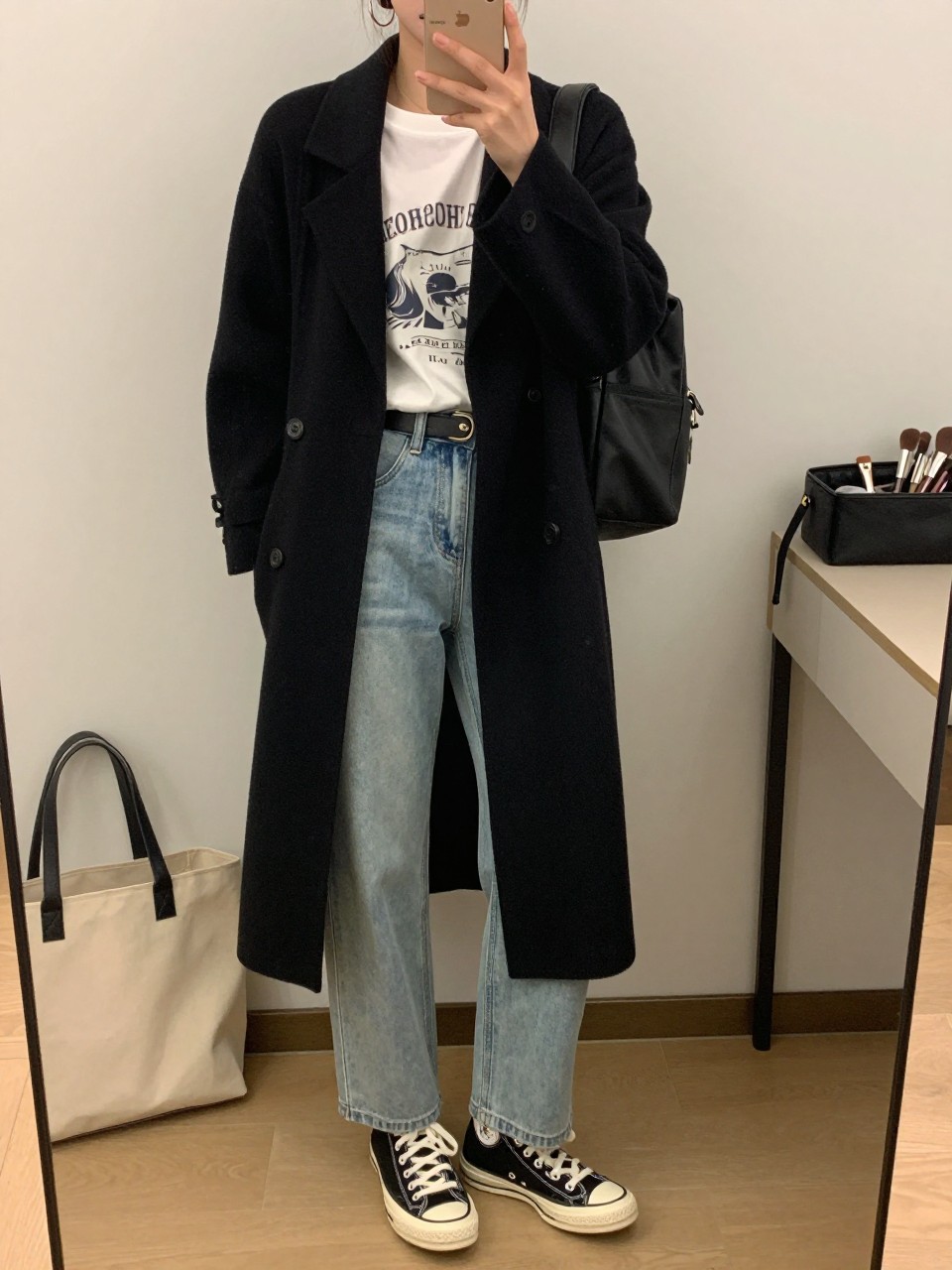 mirror selfie of a woman wearing a black long cardigan coat over a white vintage-style graphic tee, light wash straight-leg jeans, black Converse-style sneakers, silver hoop earrings, and a black backpack, clearly showing the same clothing items, colors, fabrics, and accessories listed in the Outfit Pieces section, holding a phone in front of a mirror, cropped at NECK so the face does not show, with a relaxed casual selfie pose, in a minimal dressing room, soft indoor lighting, including a tote bag resting on the floor and makeup brush or cosmetic bag on the counter, latest iPhone photo quality.