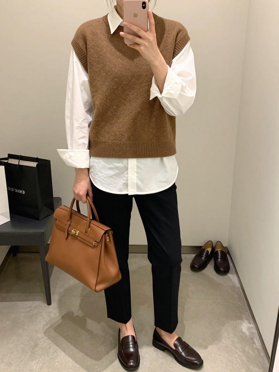 Mirror selfie of a woman wearing a mocha brown wool sweater vest layered over an oversized white poplin shirt, black tailored straight-leg trousers, dark brown leather penny loafers, and a structured cognac leather satchel, clearly showing the exact clothing items, colors, fabrics, and accessories listed, holding a latest-model iPhone in front of a mirror, cropped at the neck so the face does not show, relaxed casual selfie pose, in a minimal dressing room with soft indoor lighting, shopping bags beside a chair and a pair of loafers on the floor nearby, natural unstaged setting, latest iPhone photo quality.