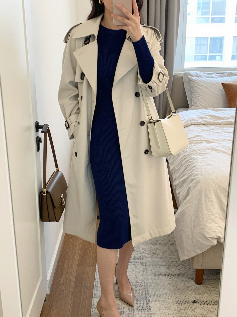 mirror selfie of a Latina woman wearing a navy ribbed knit matching top and midi skirt set, cream trench coat, nude pointed heels, gold hoop earrings, and a cream shoulder bag, holding a phone in front of a mirror, cropped at NECK so the face does not show, relaxed casual selfie pose, in a feminine-themed bedroom with natural window light, including a handbag hanging on a door handle and soft pillows on an unmade bed, realistic latest iPhone photo quality.