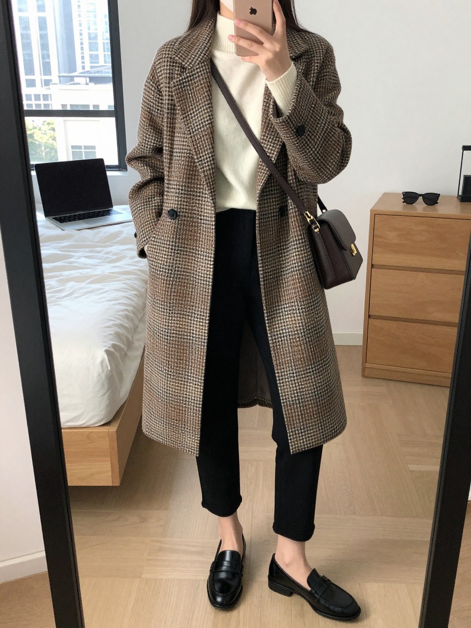 mirror selfie of a woman wearing a brown, cream, and black plaid wool coat, fitted cream cashmere sweater, black ankle-length cigarette pants, black leather loafers, and a dark brown crossbody bag, clearly showing the same clothing items, colors, fabrics, and accessories listed in the Outfit Pieces section, holding a phone in front of a mirror, cropped at NECK so the face does not show, with a relaxed casual selfie pose, in a stylish city apartment, natural window light, including a laptop on the bed and sunglasses placed on a dresser, latest iPhone photo quality.