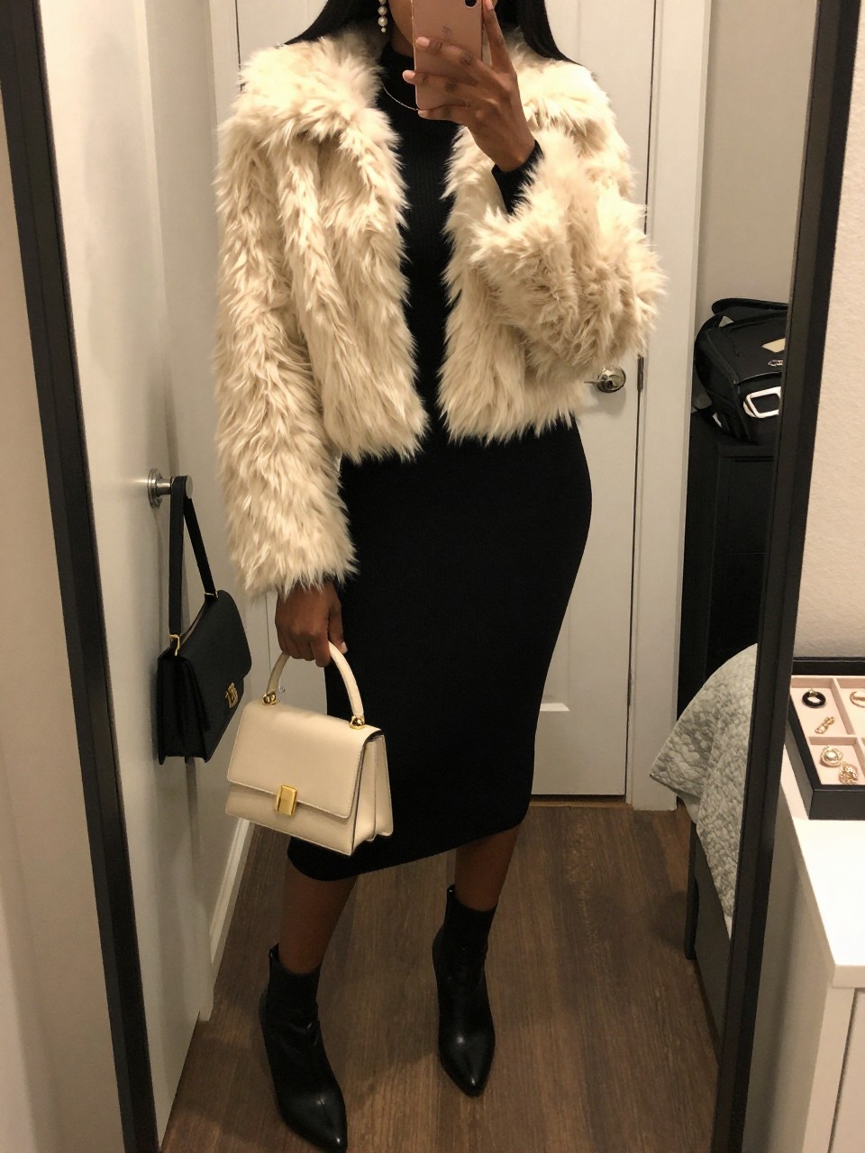 mirror selfie of a Black woman wearing the exact outfit described above, clearly showing a cream cropped faux fur jacket layered over a black long-sleeve sweater midi dress, black heeled ankle boots, a cream mini top-handle bag, and pearl drop earrings, holding a phone in front of a mirror, cropped at NECK so the face does not show, with a relaxed casual selfie pose, in a cozy bedroom mirror setup, warm moody lighting, with a handbag hanging on a door handle and a jewelry tray on the vanity, latest iPhone photo quality.