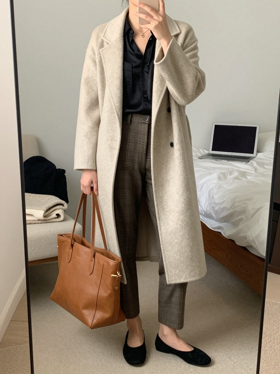 mirror selfie of a woman wearing an oatmeal long coatigan, black silk shell top, brown plaid ankle pants, black suede ballet flats, and tan leather work tote, clearly showing the same clothing items, colors, fabrics, and accessories listed in the Outfit Pieces section, holding a phone in front of a mirror, cropped at NECK so the face does not show, with a relaxed casual selfie pose, in a cozy bedroom mirror setup, soft indoor lighting, including a laptop or tablet on the bed and a folded blanket on a chair, latest iPhone photo quality.