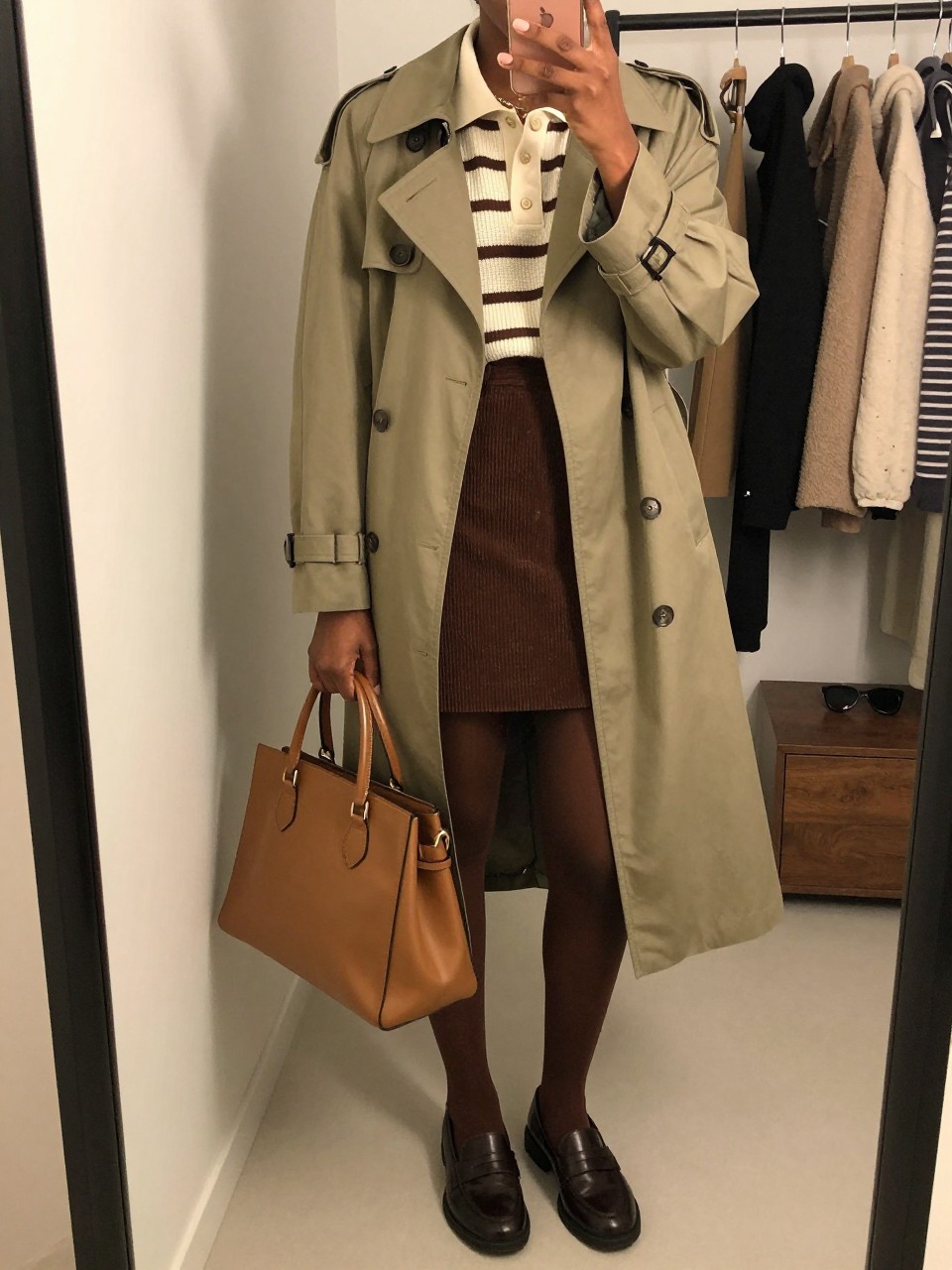 mirror selfie of a Black woman wearing a lightweight olive green trench coat over a cream and brown striped knit polo shirt, chocolate brown corduroy mini skirt, sheer brown tights, dark brown leather loafers, and a tan satchel bag, clearly showing the same clothing items, colors, fabrics, footwear, and accessories listed in the Outfit Pieces section, holding a phone in front of a mirror, cropped at NECK so the face does not show, with a relaxed casual selfie pose, in a fashion studio mirror corner, warm moody lighting, including a clothing rack with garments behind and sunglasses placed on a dresser, latest iPhone photo quality.