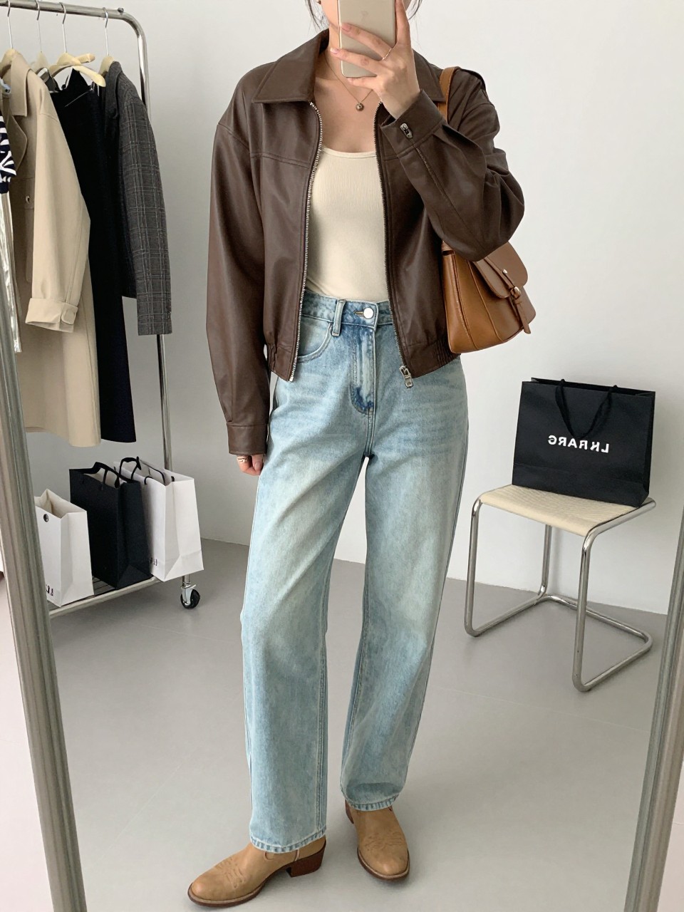 mirror selfie of a woman wearing the exact outfit described above, clearly showing a brown faux leather shacket, fitted cream bodysuit, light-wash relaxed jeans, tan western boots, and a caramel mini shoulder bag, holding a phone in front of a mirror, cropped at NECK so the face does not show, with a relaxed casual selfie pose, in a fashion studio mirror corner, natural window light, with clothing rack with garments behind and shopping bags beside a chair, latest iPhone photo quality.