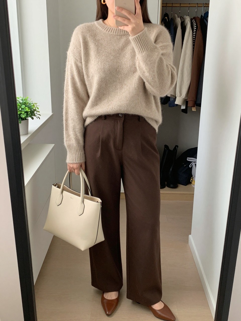 mirror selfie of a curvy woman wearing an oatmeal soft knit sweater tucked into chocolate brown wide-leg trousers, brown pointed flats, gold hoop earrings, and a cream structured tote bag, clearly showing the same clothing items, colors, fabrics, and accessories listed in the Outfit Pieces section, holding a phone in front of a mirror, cropped at NECK so the face does not show, with a relaxed casual selfie pose, in a walk-in closet mirror, natural window light, including open closet with clothes visible and a small plant on a dresser, latest iPhone photo quality.