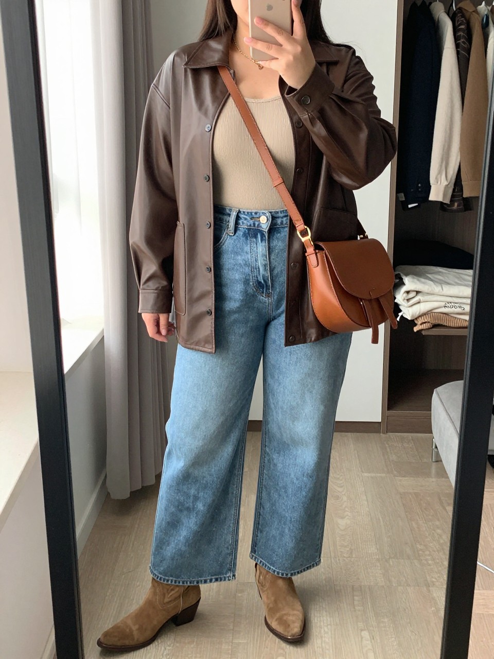 mirror selfie of a plus size woman wearing the exact outfit described above, an espresso brown faux leather shacket worn open over an oatmeal beige ribbed tank top, relaxed light-wash wide-leg jeans, tan suede western ankle boots, and cognac leather crossbody saddle bag, clearly showing the same clothing items, colors, fabrics, and accessories listed in the Outfit Pieces section, holding a phone in front of a mirror, cropped at NECK so the face does not show, with a relaxed casual selfie pose, in a walk-in closet mirror, natural window light, including an open closet with clothes visible and a folded blanket on a chair, latest iPhone photo quality.