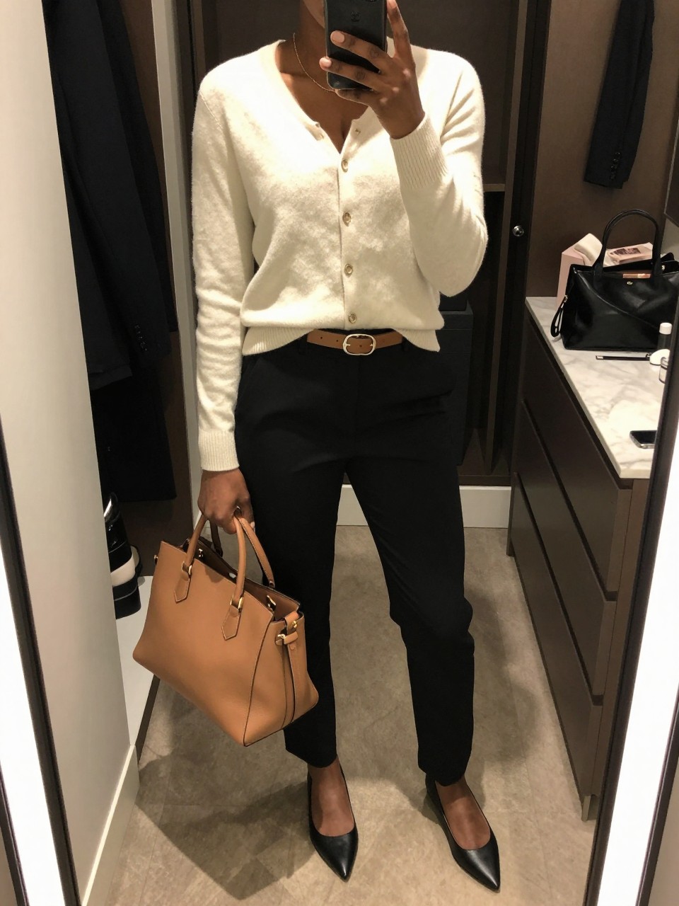 mirror selfie of a Black woman wearing a soft cream button-front cardigan, black tailored straight-leg trousers, black pointed flats, a camel leather belt, and a structured camel handbag, clearly showing the same clothing items, colors, fabrics, and accessories listed in the Outfit Pieces section, holding a phone in front of a mirror, cropped at NECK so the face does not show, with a relaxed casual selfie pose, in a walk-in closet mirror, warm moody lighting, including an open closet with clothes visible and a cosmetic bag on the counter, latest iPhone photo quality.