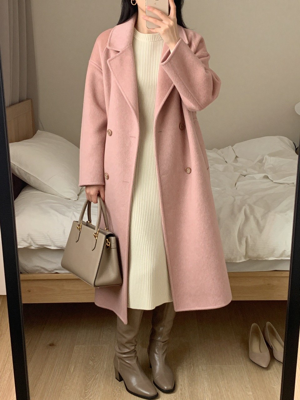 mirror selfie of a woman wearing a long blush pink wool coat over a cream ribbed knit midi dress, taupe leather knee-high boots, delicate gold hoop earrings, and a structured beige handbag, clearly showing the same clothing items, colors, fabrics, and accessories listed in the Outfit Pieces section, holding a phone in front of a mirror, cropped at NECK so the face does not show, with a relaxed casual selfie pose, in a feminine-themed bedroom, warm moody lighting, including soft pillows on an unmade bed and a pair of heels on the floor nearby, latest iPhone photo quality.
