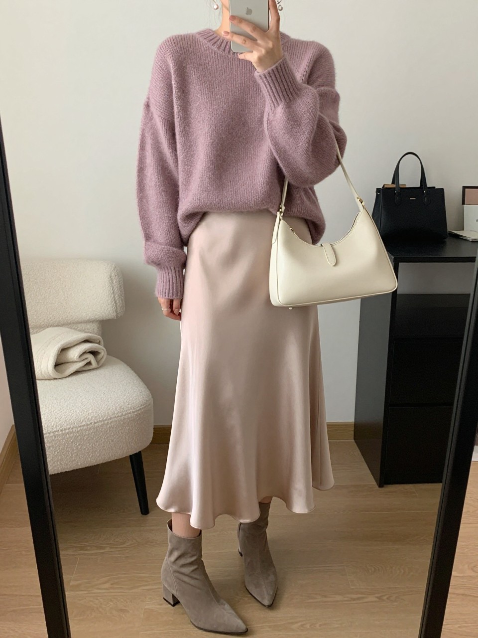 mirror selfie of a woman wearing a soft mauve oversized knit sweater, champagne satin midi skirt, taupe pointed ankle boots, small pearl drop earrings, and a cream leather shoulder bag, clearly showing the same clothing items, colors, fabrics, and accessories listed in the Outfit Pieces section, holding a phone in front of a mirror, cropped at NECK so the face does not show, with a relaxed casual selfie pose, in a cozy bedroom mirror setup, soft indoor lighting, including a folded blanket on a chair and a cosmetic bag on the counter, latest iPhone photo quality.