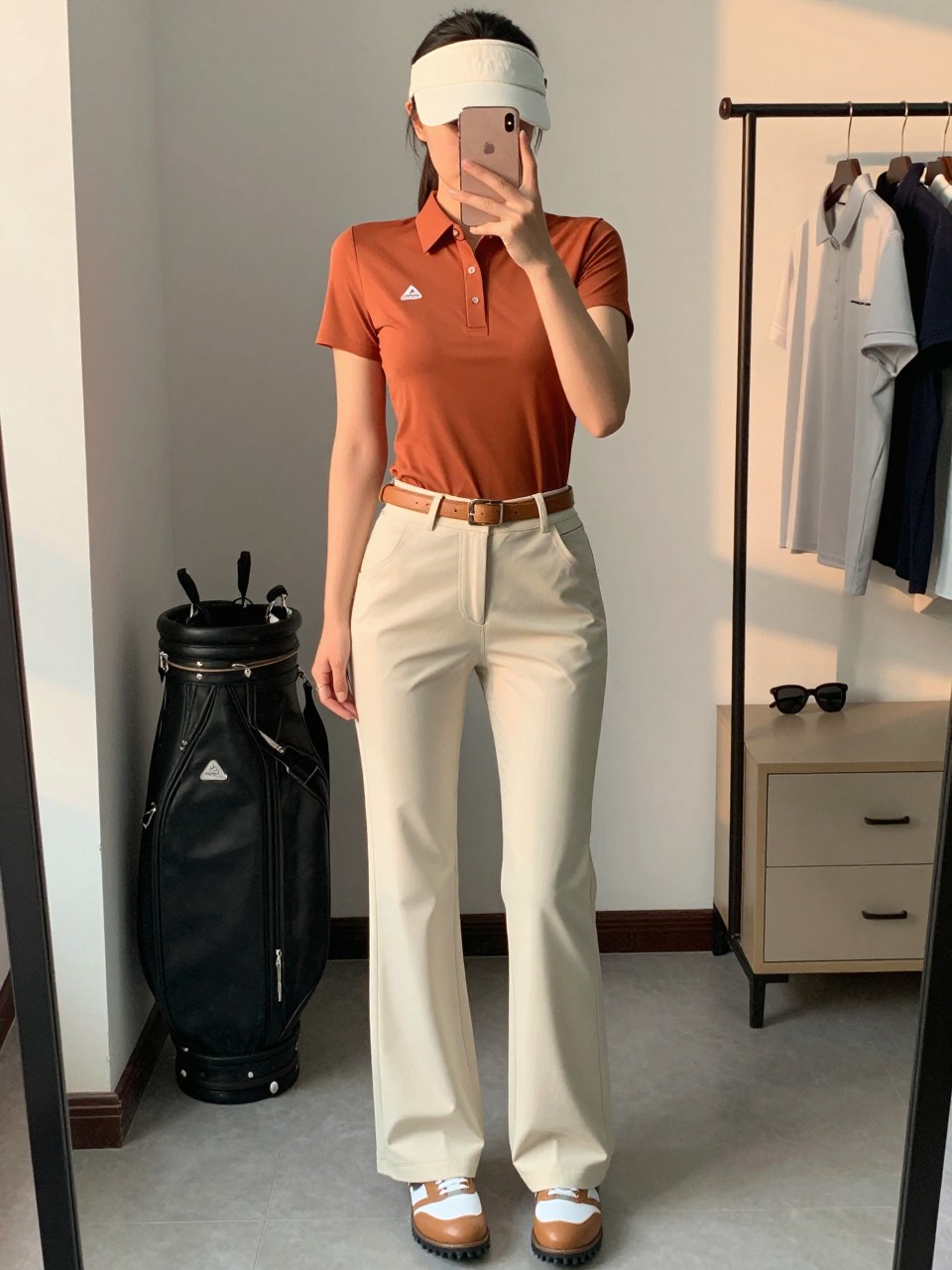 Mirror selfie of a woman wearing a fitted copper-orange short-sleeve performance golf polo tucked into cream high-waisted wide-leg golf pants made from flexible lightweight fabric, tan-and-white spikeless golf shoes, a slim tan leather belt, and a cream visor, clearly showing the exact clothing items, colors, fabrics, and accessories listed, holding a latest-model iPhone in front of a mirror, cropped at the neck so the face does not show, relaxed casual selfie pose, in a fashion studio mirror corner with evening golden-hour lighting, a small golf tote beside a clothing rack and sunglasses resting on a dresser, natural unstaged composition, latest iPhone photo quality.