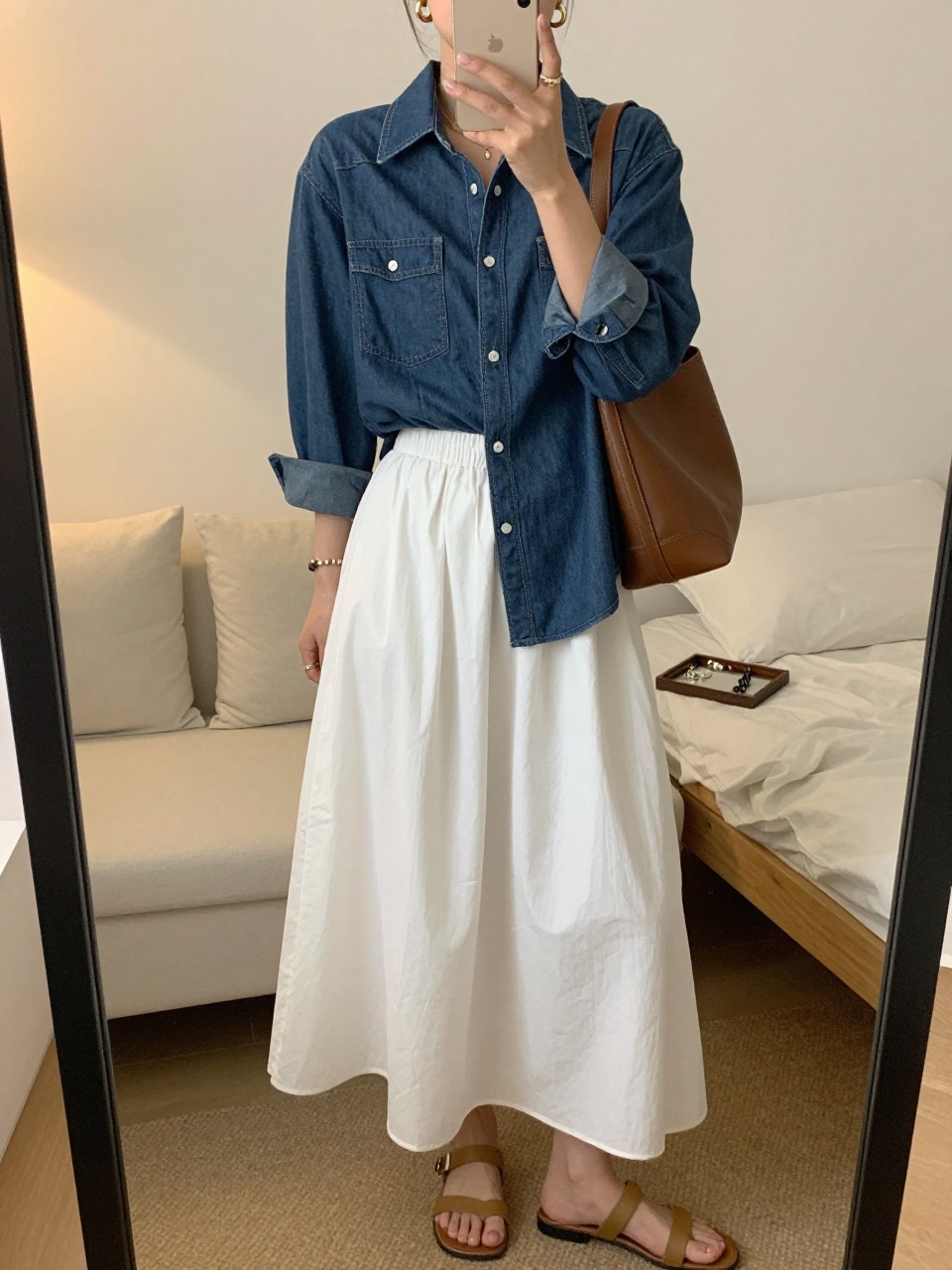 mirror selfie of a woman wearing a medium-wash denim button-down shirt, white cotton maxi skirt, tan flat sandals, a brown leather shoulder bag, and gold hoop earrings, clearly showing the same clothing items, colors, fabrics, and accessories listed in the Outfit Pieces section, holding a phone in front of a mirror, cropped at NECK so the face does not show, with a relaxed casual selfie pose, in a feminine-themed bedroom, evening golden hour, including soft pillows on an unmade bed and a jewelry tray on the vanity, latest iPhone photo quality.