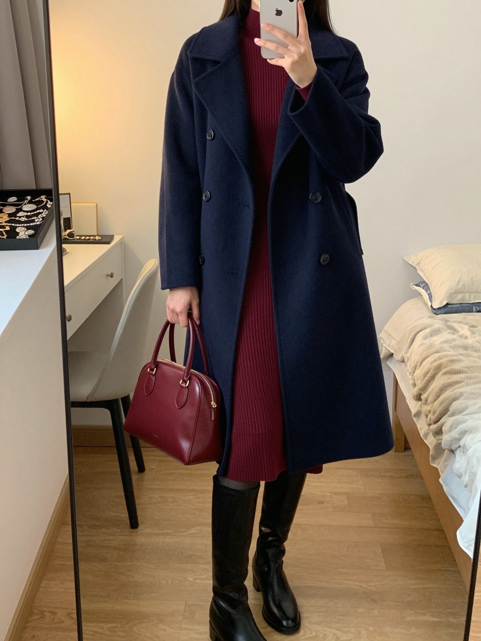 Mirror selfie of a woman wearing a knee-length navy wool funnel-neck coat over a fitted burgundy ribbed-knit midi dress, black opaque tights, knee-high black leather boots, and a compact burgundy leather handbag, clearly showing the exact clothing items, colors, textures, and accessories listed, holding a latest-model iPhone in front of a mirror, cropped at the neck so the face does not show, relaxed casual selfie pose, in a feminine-themed bedroom with evening golden-hour lighting, a jewelry tray on the vanity and a folded blanket on a chair, natural unstaged composition, latest iPhone photo quality.