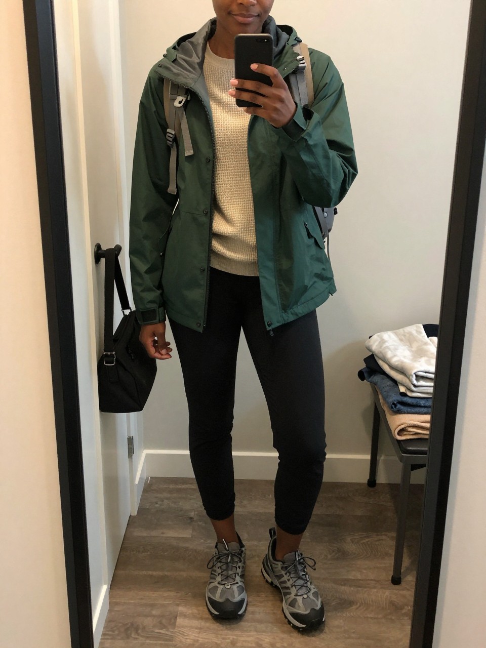 mirror selfie of a Black woman wearing the exact outfit described above, clearly showing a forest green lightweight rain jacket, oatmeal waffle-knit base layer, black tapered jogger pants, gray waterproof trail sneakers, and a beige hiking backpack, holding a phone in front of a mirror, cropped at NECK so the face does not show, with a relaxed casual selfie pose, in a walk-in closet mirror, evening golden hour, with a handbag hanging on a door handle and folded blanket on a chair, latest iPhone photo quality.