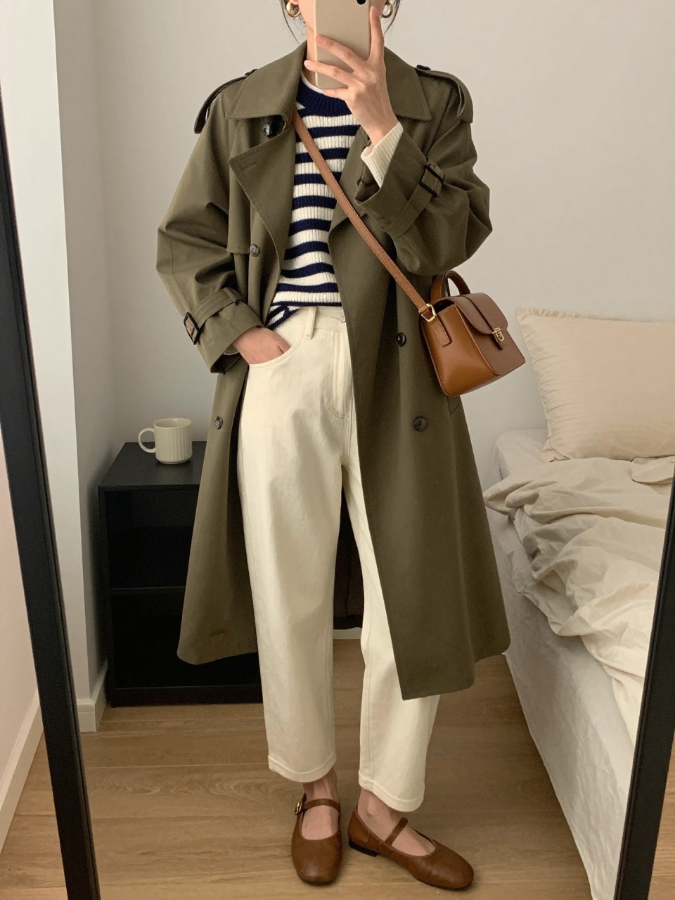 mirror selfie of a woman wearing the exact outfit described above, clearly showing an olive trench coat layered over a navy and cream striped sweater, cream barrel jeans, brown ballet flats, a tan crossbody bag, and gold hoop earrings, holding a phone in front of a mirror, cropped at NECK so the face does not show, with a relaxed casual selfie pose, in a cozy bedroom mirror setup, soft indoor lighting, with a coffee mug on a bedside table and soft pillows on an unmade bed, latest iPhone photo quality.
