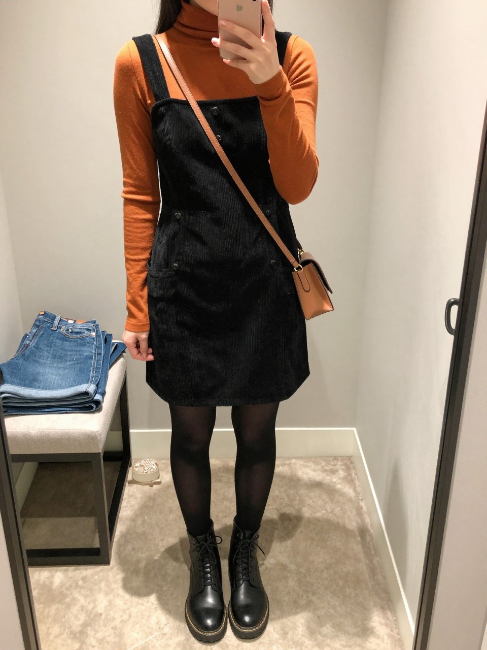 mirror selfie of a woman wearing the exact outfit described above, clearly showing a black corduroy pinafore dress, pumpkin orange fitted turtleneck, sheer black tights, black lace-up boots, and a brown mini crossbody bag, holding a phone in front of a mirror, cropped at NECK so the face does not show, with a relaxed casual selfie pose, in a walk-in closet mirror, soft indoor lighting, with folded denim jeans on a bench and a jewelry tray on the vanity, latest iPhone photo quality.