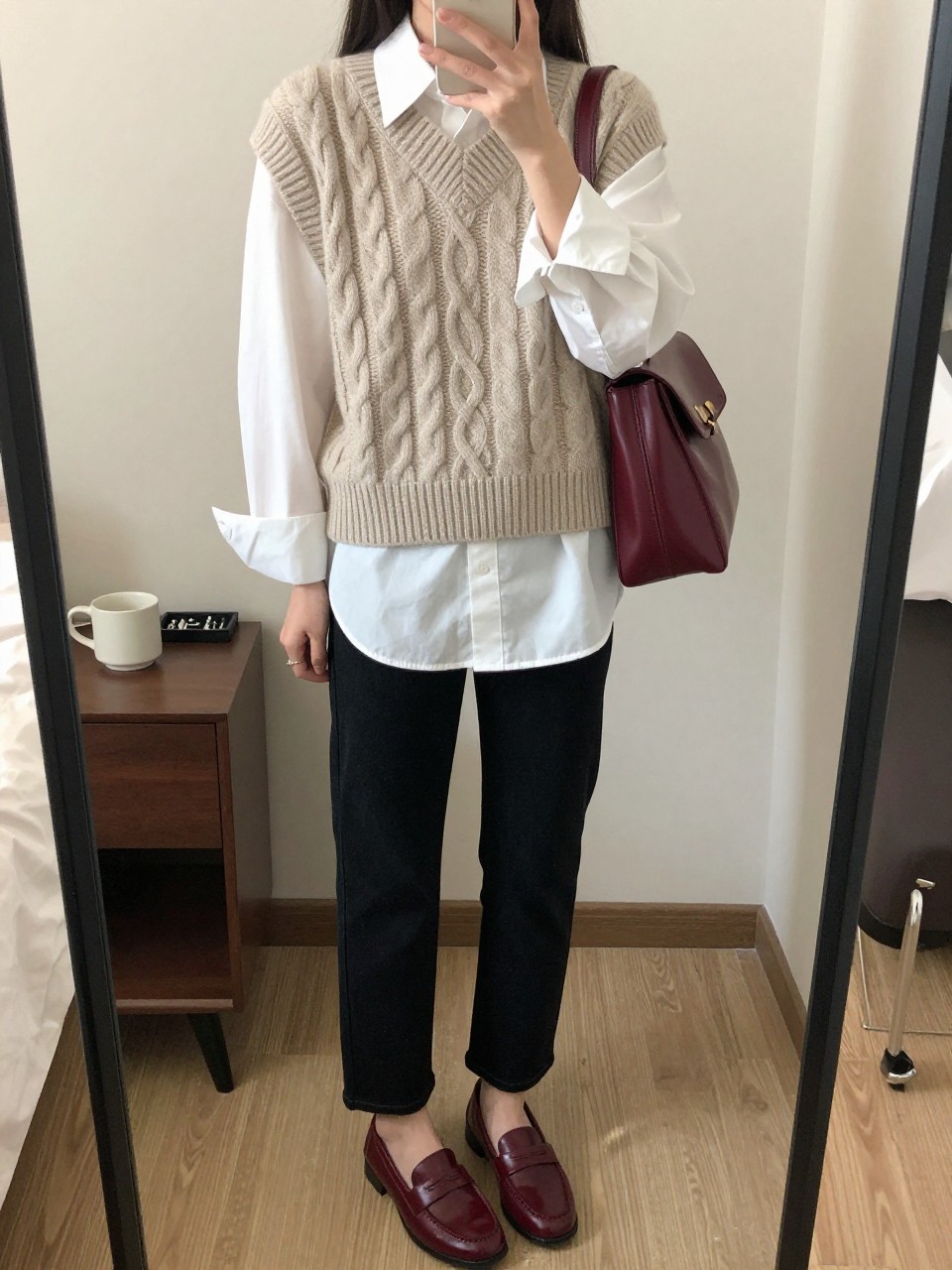 mirror selfie of a woman wearing an oatmeal cable-knit sweater vest over an oversized white button-down shirt, black straight-leg jeans, burgundy loafers, and a burgundy leather book bag, clearly showing the same clothing items, colors, fabrics, and accessories listed in the Outfit Pieces section, holding a phone in front of a mirror, cropped at NECK so the face does not show, with a relaxed casual selfie pose, in a cozy bedroom mirror setup, soft indoor lighting, including a coffee mug on a bedside table and a jewelry tray on the vanity, latest iPhone photo quality.