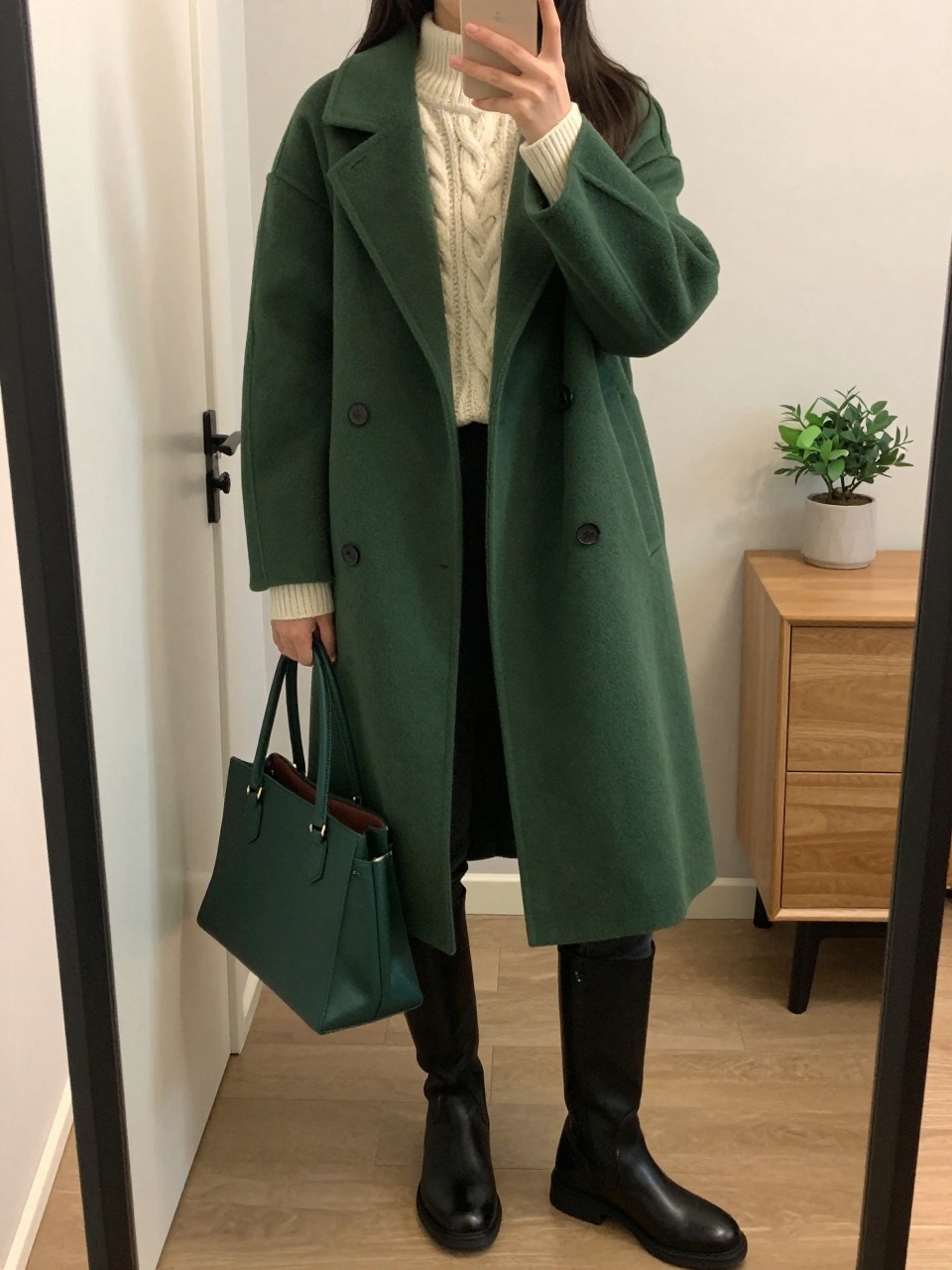 mirror selfie of a woman wearing a forest green wool coat over a cream cable-knit sweater, black skinny jeans, black riding boots, and a dark green leather handbag, clearly showing the same clothing items, colors, fabrics, and accessories listed in the Outfit Pieces section, holding a phone in front of a mirror, cropped at NECK so the face does not show, with a relaxed casual selfie pose, in a cozy bedroom mirror setup, warm moody lighting, including a handbag hanging on a door handle and a small plant on a dresser, latest iPhone photo quality.