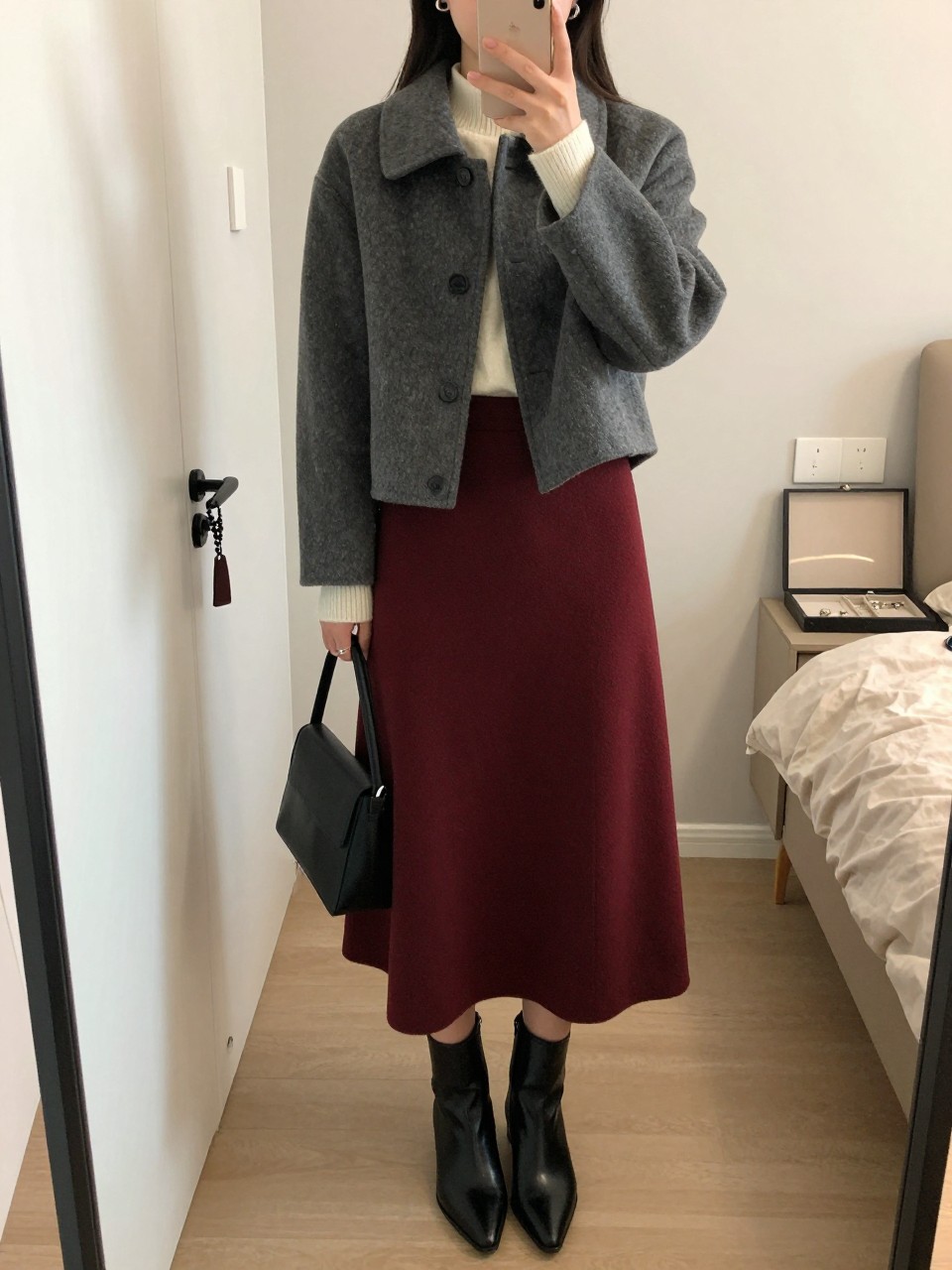 mirror selfie of a woman wearing the exact outfit described above, clearly showing a charcoal cropped wool jacket layered over an ivory mock-neck sweater, burgundy pleated midi skirt, black pointed ankle boots, a black top-handle bag, and silver hoop earrings, holding a phone in front of a mirror, cropped at NECK so the face does not show, with a relaxed casual selfie pose, in a feminine-themed bedroom, soft indoor lighting, with a handbag hanging on a door handle and a jewelry tray on the vanity, latest iPhone photo quality.