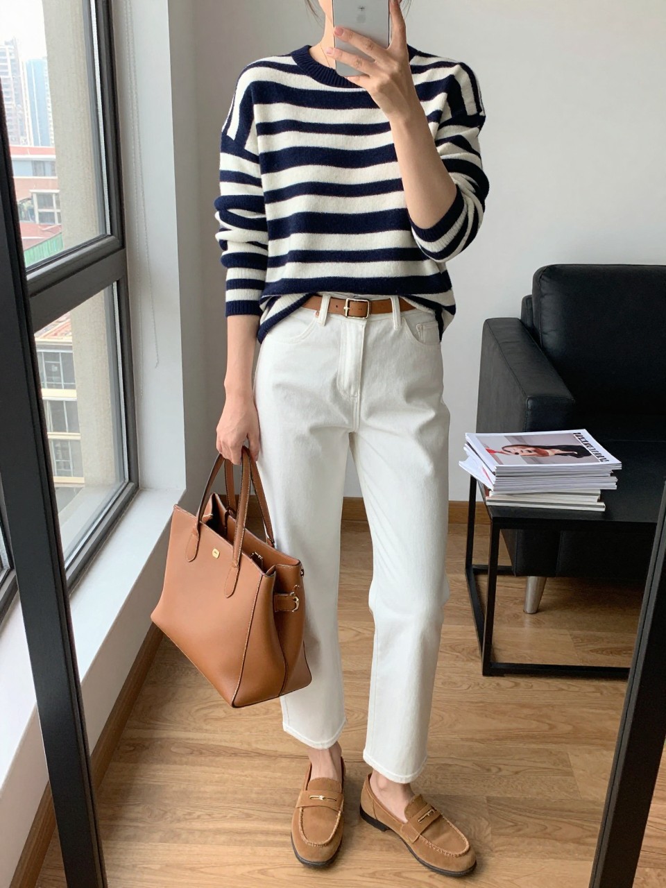 mirror selfie of a woman wearing a navy and cream striped lightweight sweater, white straight-leg jeans, tan suede loafers, a camel belt, and a tan leather tote bag, clearly showing the same clothing items, colors, fabrics, and accessories listed in the Outfit Pieces section, holding a phone in front of a mirror, cropped at NECK so the face does not show, with a relaxed casual selfie pose, in a stylish city apartment, natural window light, including a tote bag resting on the floor and a stack of fashion magazines on a side table, latest iPhone photo quality.