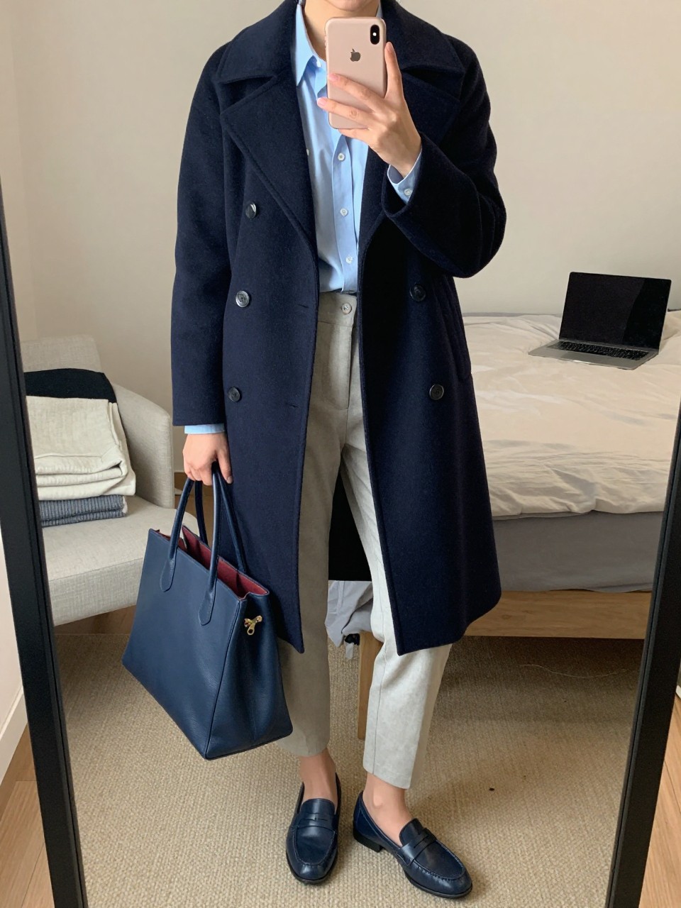 Mirror selfie of a woman wearing a tailored navy wool peacoat over a pale blue button-down shirt, stone-colored ankle-length cigarette pants, navy leather loafers, and a structured navy leather tote bag, clearly showing the exact clothing items, colors, fabrics, and accessories listed, holding a latest-model iPhone in front of a mirror, cropped at the neck so the face does not show, relaxed casual selfie pose, in a cozy bedroom mirror setup with soft indoor lighting, a laptop on the bed and folded blanket on a chair, natural unstaged composition, latest iPhone photo quality.