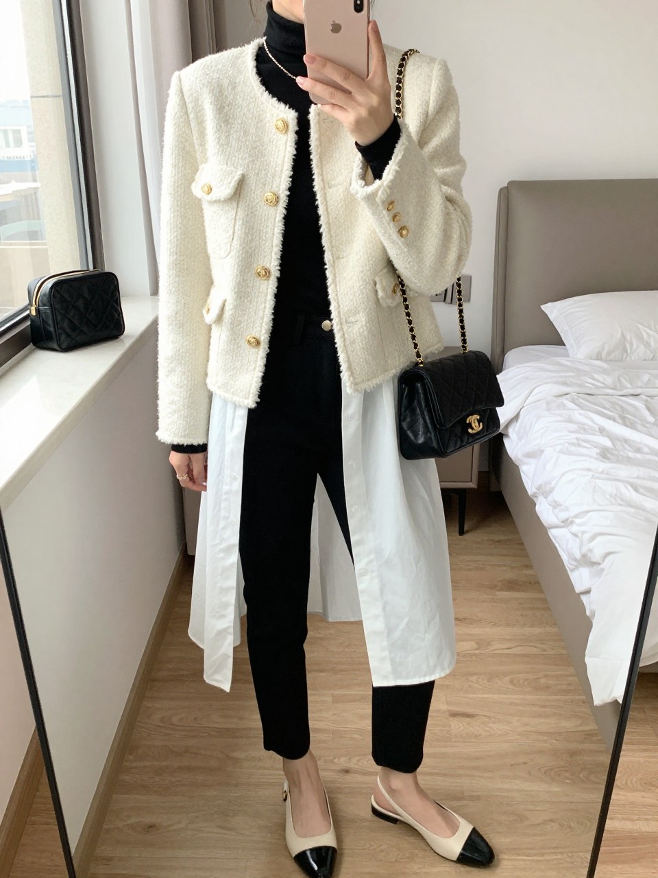 Mirror selfie of a woman wearing a cream bouclé jacket with gold buttons over a fitted black turtleneck and a long white poplin shirt with the hem showing underneath, black high-waisted slim trousers, cream-and-black slingback flats, and a quilted black shoulder bag with a gold chain, clearly showing the exact clothing items, colors, textures, and accessories listed, holding a latest-model iPhone in front of a mirror, cropped at the neck so the face does not show, relaxed casual selfie pose, in a feminine-themed bedroom with natural window light, a cosmetic bag on the counter and a folded blanket on a chair, natural casual setting, latest iPhone photo quality.