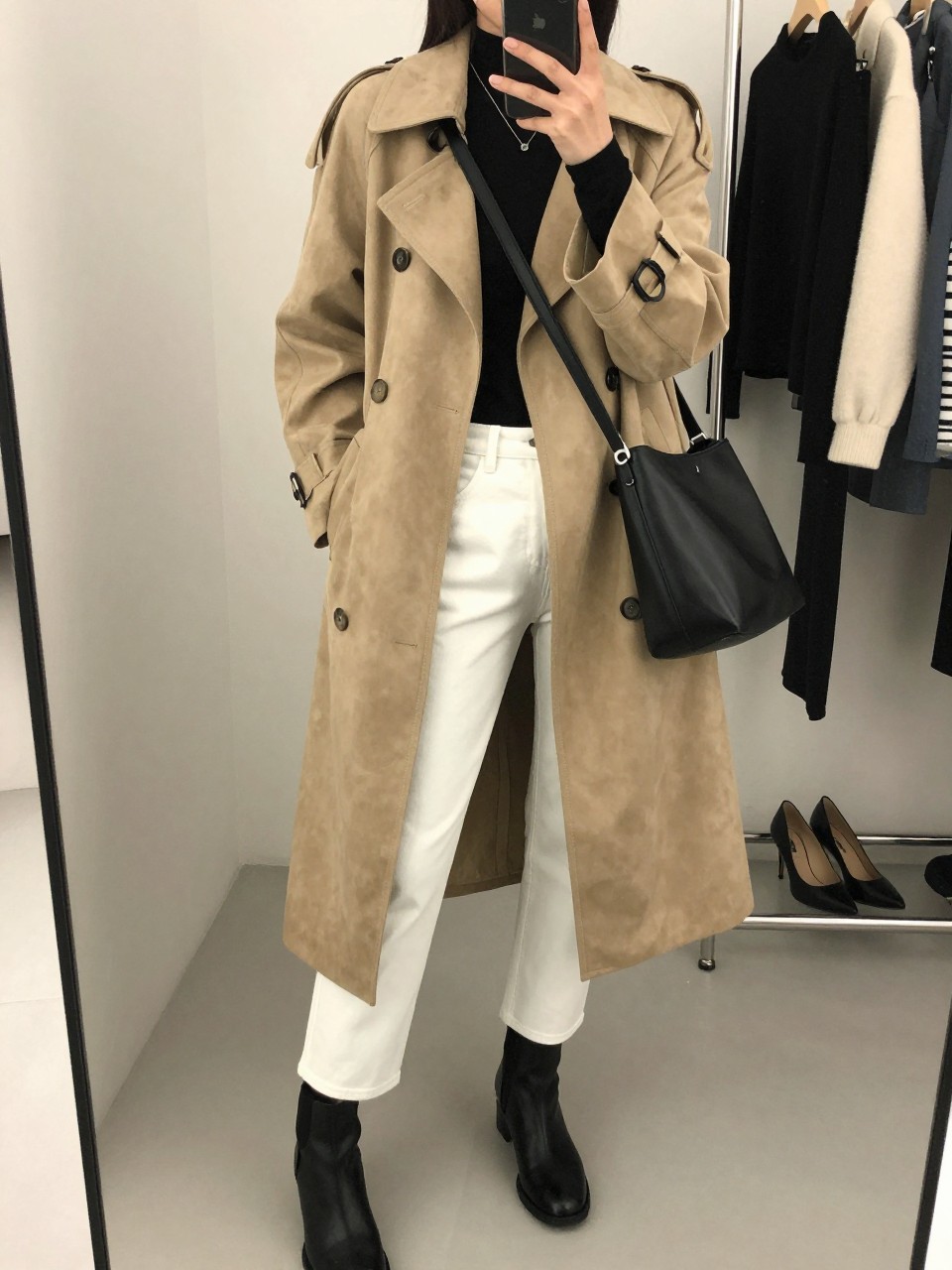 mirror selfie of a woman wearing a beige suede trench coat, fitted black long-sleeve top, straight white jeans, black ankle boots, and a black leather crossbody bag, clearly showing the same clothing items, colors, fabrics, and accessories listed in the Outfit Pieces section, holding a phone in front of a mirror, cropped at NECK so the face does not show, with a relaxed casual selfie pose, in a fashion studio mirror corner, soft indoor lighting, including a clothing rack with garments behind and a pair of heels on the floor nearby, latest iPhone photo quality.