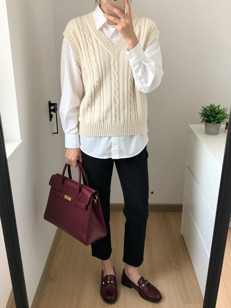 mirror selfie of a woman wearing a cream cable-knit sweater vest over a white poplin button-down shirt, black ankle pants, burgundy loafers, and a burgundy leather satchel, clearly showing the same clothing items, colors, fabrics, and accessories listed in the Outfit Pieces section, holding a phone in front of a mirror, cropped at NECK so the face does not show, with a relaxed casual selfie pose, in a walk-in closet mirror, natural window light, including a handbag hanging on a door handle and a small plant on a dresser, latest iPhone photo quality.