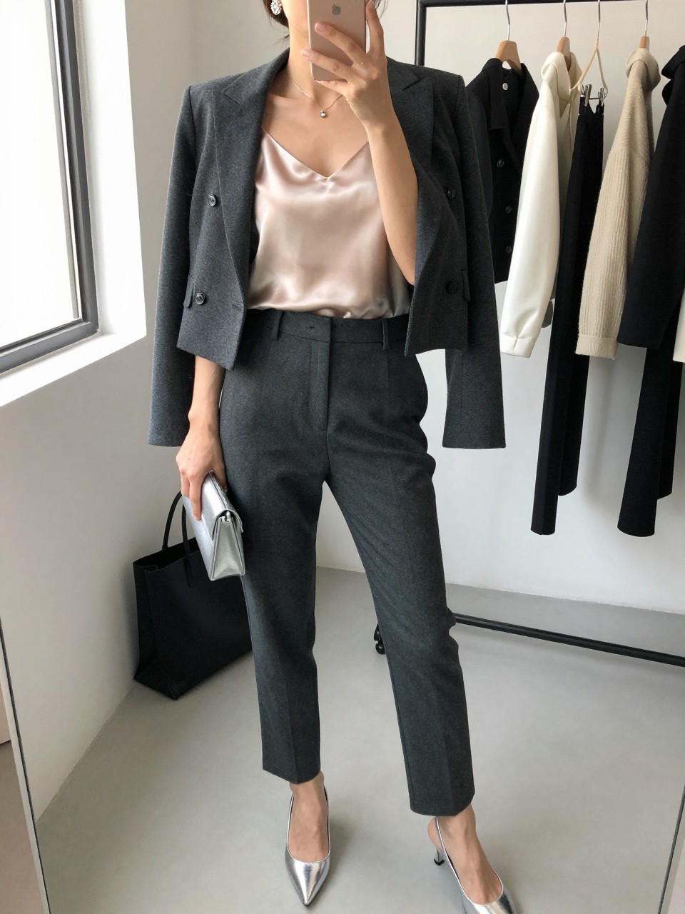 mirror selfie of a woman wearing the exact outfit described above, clearly showing a champagne satin cami layered under a charcoal cropped blazer, charcoal tailored trousers, silver pointed heels, a silver clutch, and crystal stud earrings, holding a phone in front of a mirror, cropped at NECK so the face does not show, with a relaxed casual selfie pose, in a fashion studio mirror corner, natural window light, with clothing rack with garments behind and a tote bag resting on the floor, latest iPhone photo quality.