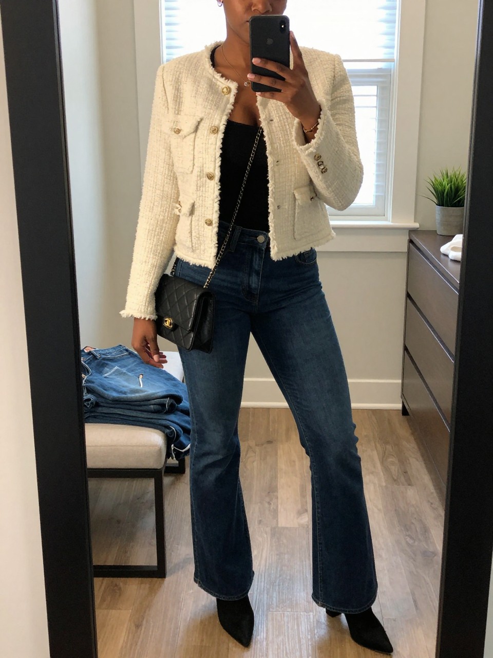mirror selfie of a Black woman wearing the exact outfit described above, clearly showing a cream tweed jacket, black fitted tank, dark blue flare jeans, black heeled boots, and a quilted black mini bag, holding a phone in front of a mirror, cropped at NECK so the face does not show, with a relaxed casual selfie pose, in a walk-in closet mirror, natural window light, with folded denim jeans on a bench and a small plant on a dresser, latest iPhone photo quality.