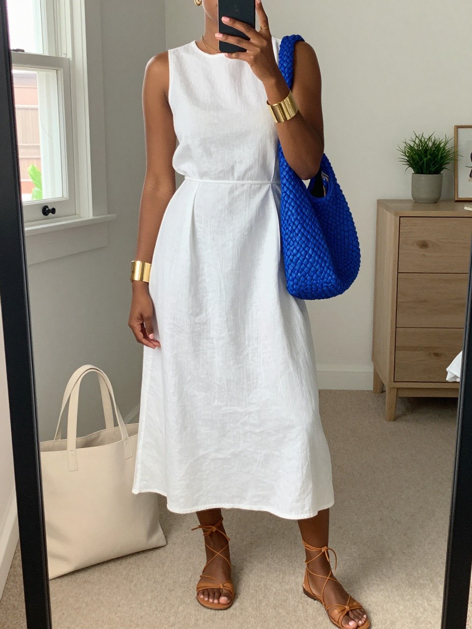 mirror selfie of a Black woman wearing a white sleeveless linen midi dress with a relaxed fitted waist, tan lace-up flat sandals, cobalt blue woven shoulder bag, gold cuff bracelet, and small gold hoop earrings, clearly showing the same clothing items, colors, fabrics, and accessories listed in the Outfit Pieces section, holding a phone in front of a mirror, cropped at NECK so the face does not show, with a relaxed casual selfie pose, in a feminine-themed bedroom, natural window light, including a cream tote bag resting on the floor and a small plant on a dresser, latest iPhone photo quality.