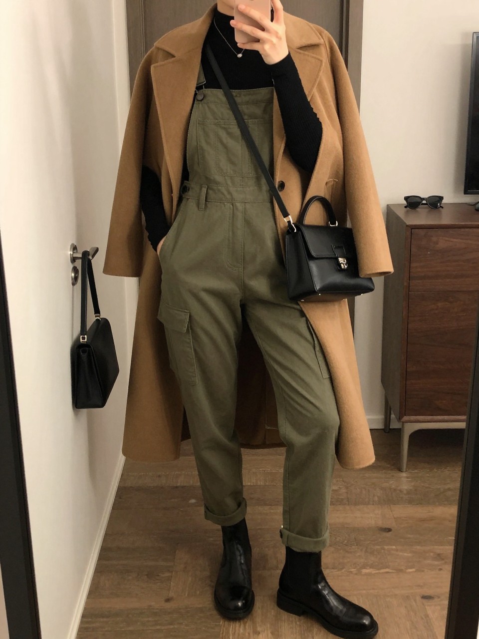 mirror selfie of a woman wearing olive green tapered utility overalls over a fitted black ribbed long-sleeve top, layered beneath a long camel wool coat, black leather Chelsea boots, and a structured black crossbody bag, clearly showing the same clothing items, colors, fabrics, footwear, and accessories listed in the Outfit Pieces section, holding a phone in front of a mirror, cropped at NECK so the face does not show, with a relaxed casual selfie pose, in a stylish city apartment, warm moody lighting, including a handbag hanging on a door handle and sunglasses placed on a dresser, latest iPhone photo quality.