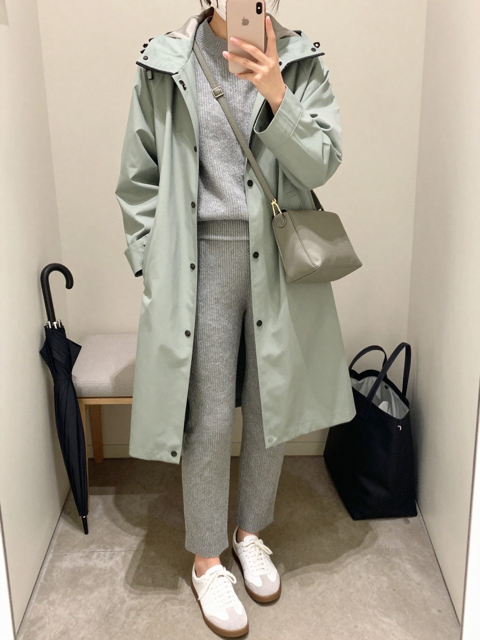 Mirror selfie of a woman wearing a knee-length sage green waterproof raincoat over a matching heather gray ribbed-knit sweater and straight-leg knit pants set, white leather sneakers with gray suede details, and a compact sage nylon crossbody bag, clearly showing the exact clothing items, colors, fabrics, and accessories listed, holding a latest-model iPhone in front of a mirror, cropped at the neck so the face does not show, relaxed casual selfie pose, in a minimal dressing room with soft indoor lighting, an umbrella leaning beside a bench and a tote bag resting on the floor, natural uncluttered composition, latest iPhone photo quality.