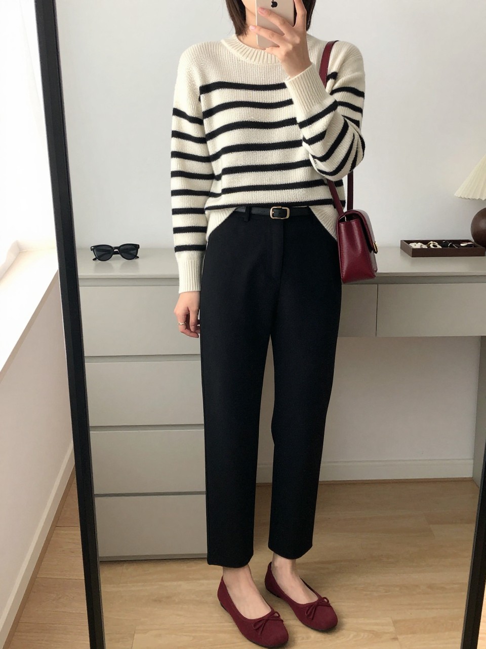 mirror selfie of a woman wearing a cream and black striped knit sweater, black tailored straight-leg trousers, burgundy ballet flats, a slim black belt, and a small burgundy shoulder bag, clearly showing the same clothing items, colors, fabrics, and accessories listed in the Outfit Pieces section, holding a phone in front of a mirror, cropped at NECK so the face does not show, with a relaxed casual selfie pose, in a minimal dressing room, natural window light, including sunglasses placed on a dresser and a jewelry tray on the vanity, latest iPhone photo quality.