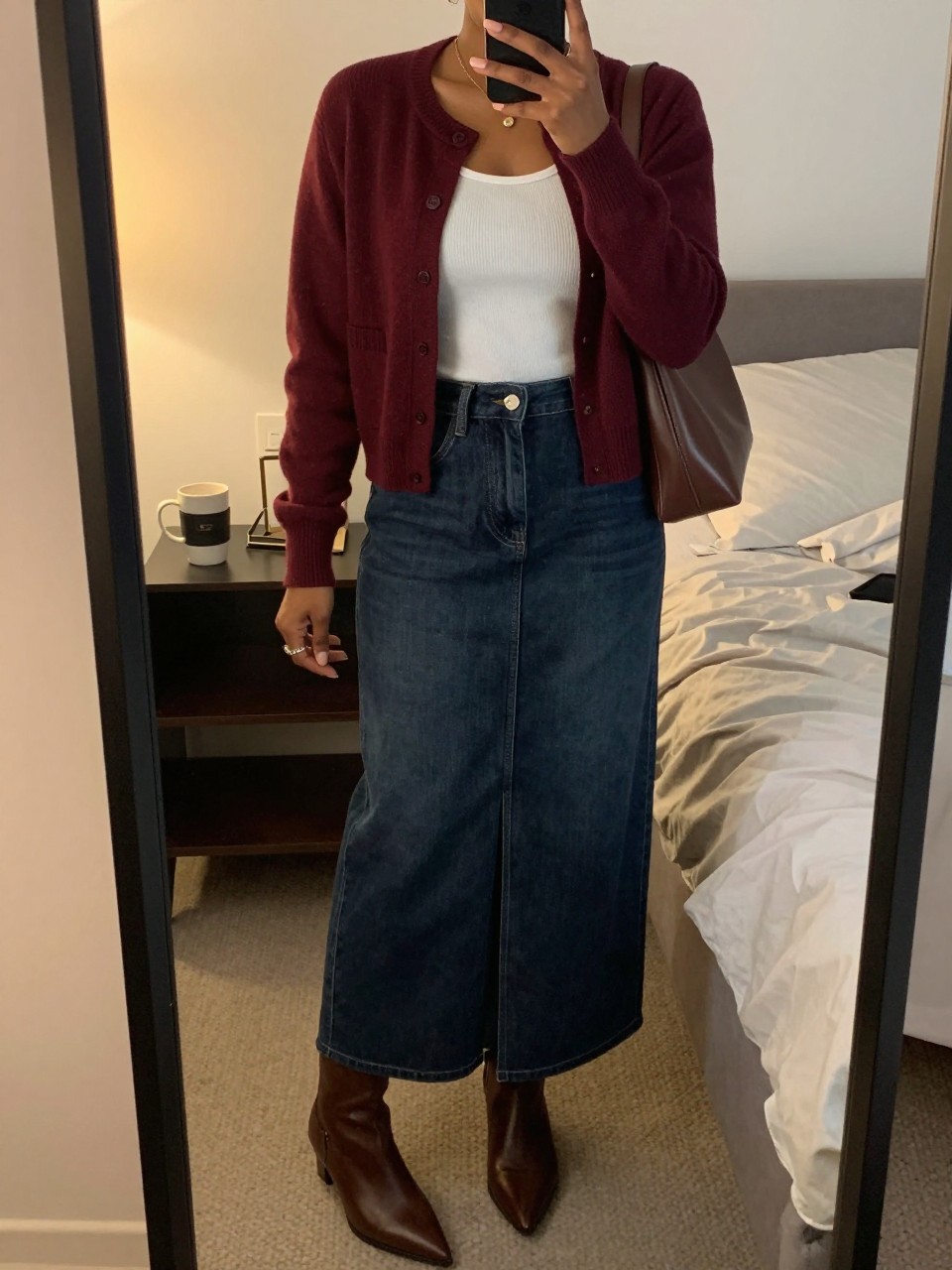mirror selfie of a Black woman wearing a burgundy button-front cardigan, white fitted ribbed tank top, dark-wash denim maxi skirt with a front slit, brown pointed ankle boots, and a chocolate brown shoulder bag, clearly showing the same clothing items, colors, fabrics, and accessories listed in the Outfit Pieces section, holding a phone in front of a mirror, cropped at NECK so the face does not show, with a relaxed casual selfie pose, in a cozy bedroom mirror setup, evening golden hour, including a coffee mug on a bedside table and soft pillows on an unmade bed, latest iPhone photo quality.