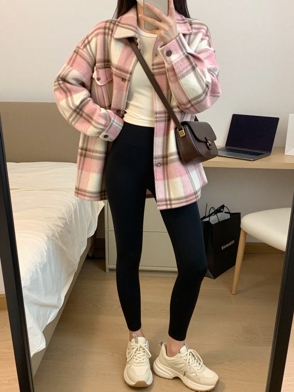mirror selfie of a woman wearing a pink, cream, and brown plaid oversized shacket over a fitted cream long-sleeve top, matte black high-waisted leggings, chunky cream sneakers, and a chocolate brown belt bag worn crossbody, clearly showing the same clothing items, colors, fabrics, and accessories listed in the Outfit Pieces section, holding a phone in front of a mirror, cropped at NECK so the face does not show, with a relaxed casual selfie pose, in a cozy bedroom mirror setup, soft indoor lighting, including a laptop on the bed and shopping bags beside a chair, latest iPhone photo quality.