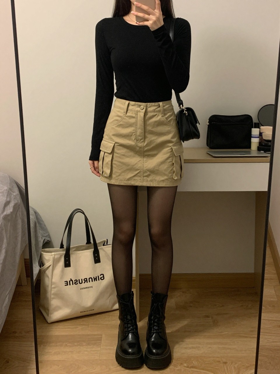mirror selfie of a woman wearing a fitted black graphic long-sleeve tee, khaki cargo mini skirt, sheer black tights, black platform combat boots, and a small black shoulder bag, clearly showing the same clothing items, colors, fabrics, and accessories listed in the Outfit Pieces section, holding a phone in front of a mirror, cropped at NECK so the face does not show, with a relaxed casual selfie pose, in a cozy bedroom mirror setup, warm moody lighting, including a tote bag resting on the floor and a cosmetic bag on the counter, latest iPhone photo quality.