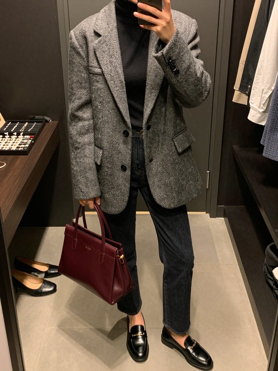 mirror selfie of a woman wearing an oversized gray herringbone wool blazer, fitted black turtleneck, dark-wash high-waisted straight-leg jeans, black leather penny loafers, and a structured burgundy top-handle bag, clearly showing the same clothing items, colors, fabrics, footwear, and accessories listed in the Outfit Pieces section, holding a phone in front of a mirror, cropped at NECK so the face does not show, with a relaxed casual selfie pose, in a walk-in closet mirror, warm moody lighting, including a jewelry tray on the vanity and a pair of heels on the floor nearby, latest iPhone photo quality.