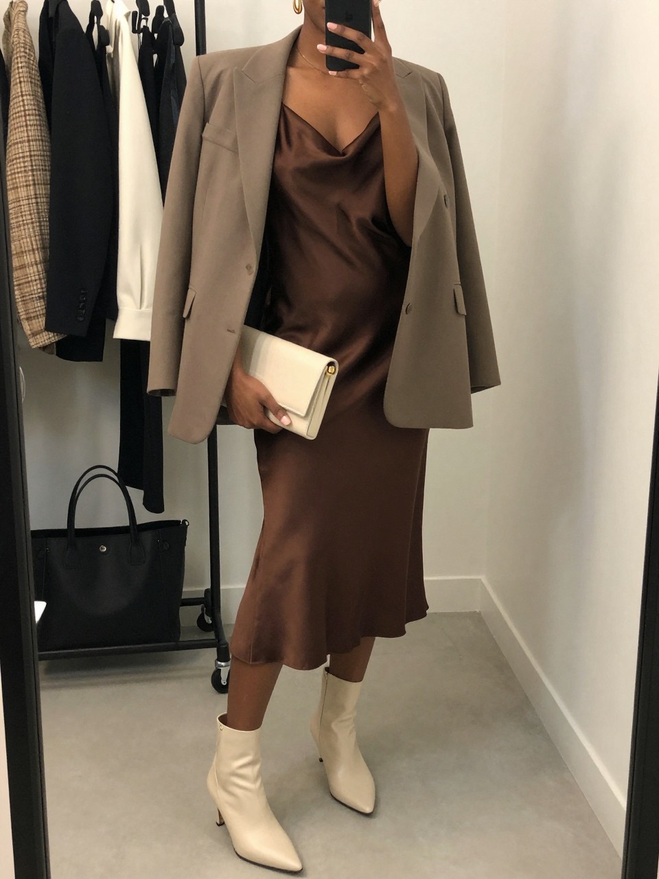 mirror selfie of a Black woman wearing the exact outfit described above, clearly showing a chocolate brown satin midi slip dress, taupe oversized blazer, cream kitten-heel ankle boots, a cream clutch, and gold drop earrings, holding a phone in front of a mirror, cropped at NECK so the face does not show, with a relaxed casual selfie pose, in a fashion studio mirror corner, soft indoor lighting, with clothing rack with garments behind and a tote bag resting on the floor, latest iPhone photo quality.