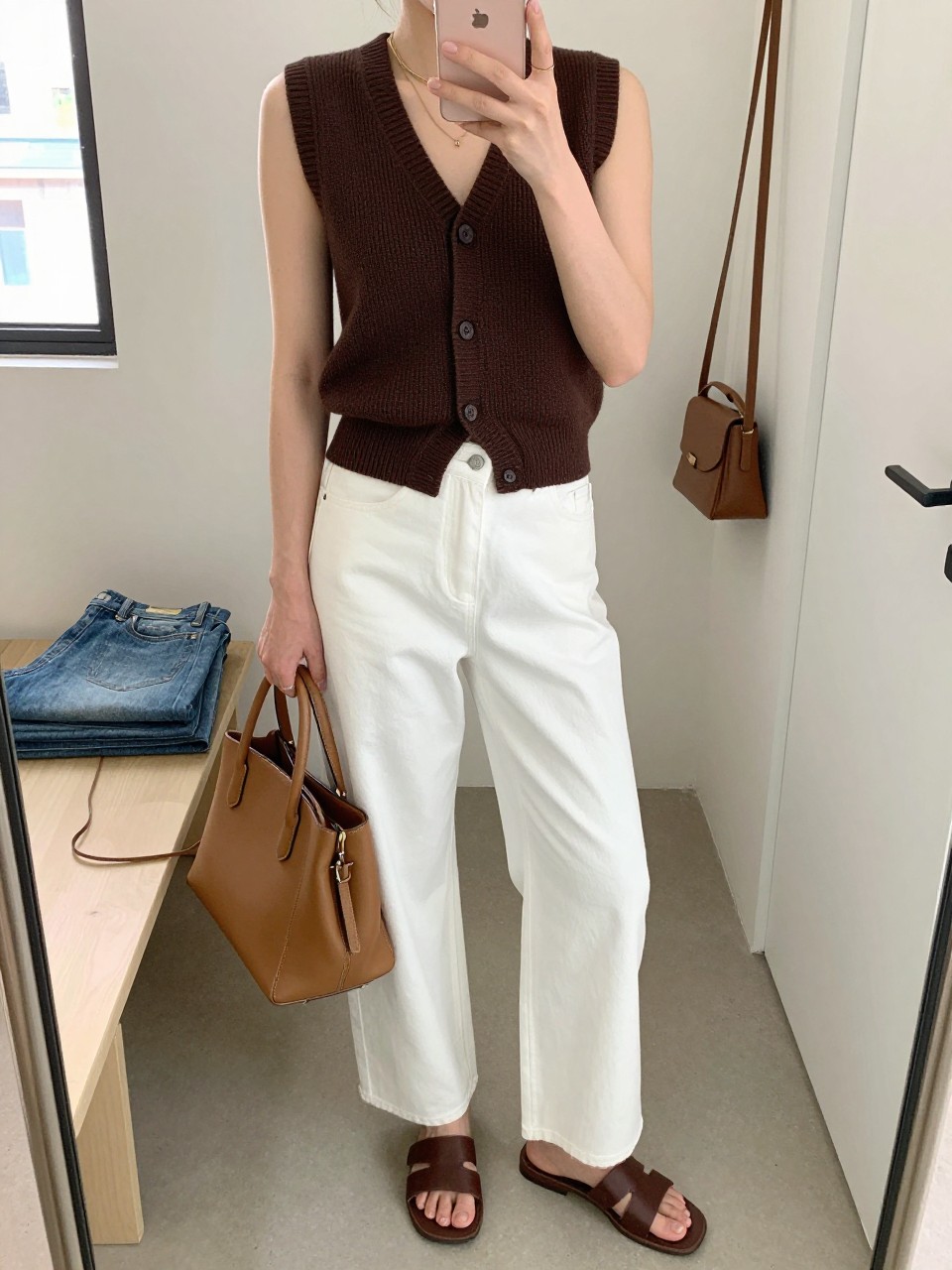 Mirror selfie of a woman wearing a sleeveless chocolate brown fine-knit vest over white high-waisted wide-leg jeans, dark brown leather slide sandals, a structured tan top-handle bag, and delicate gold layered necklaces, clearly showing the exact clothing items, colors, fabrics, and accessories listed, holding a latest-model iPhone in front of a mirror, cropped at the neck so the face does not show, relaxed casual selfie pose, in a walk-in closet mirror with natural window light, folded denim jeans on a bench and a handbag hanging on a door handle, realistic uncluttered setting, latest iPhone photo quality.